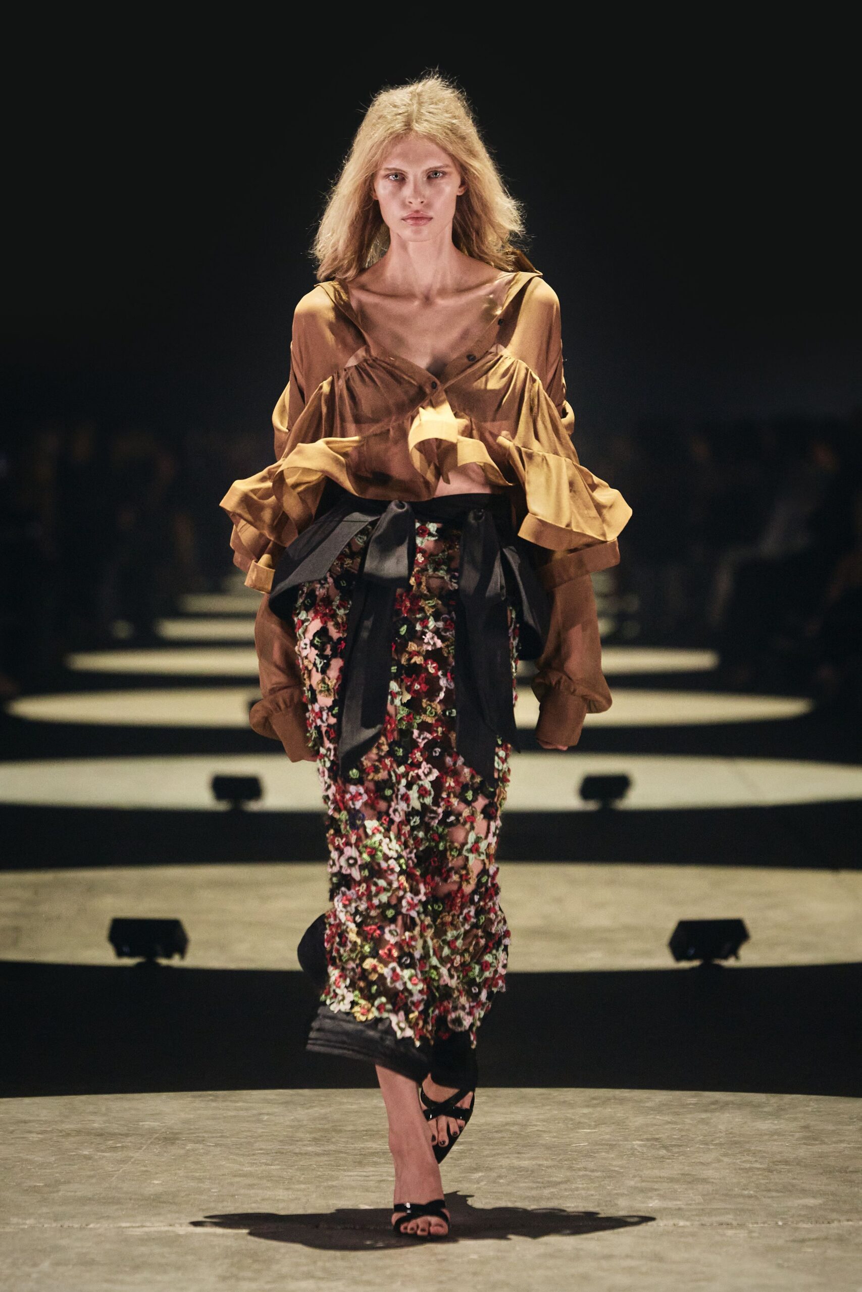 Sheer bronze blouse with dropped shoulder slashes and ruffled cuffs. Wide black sash tied in a low, floppy bow. Column skirt crusted with dense pink and burgundy floral paillettes. Black satin sandals with narrow crossed straps. For arriving at a wedding after the garden has gone damp and dark.