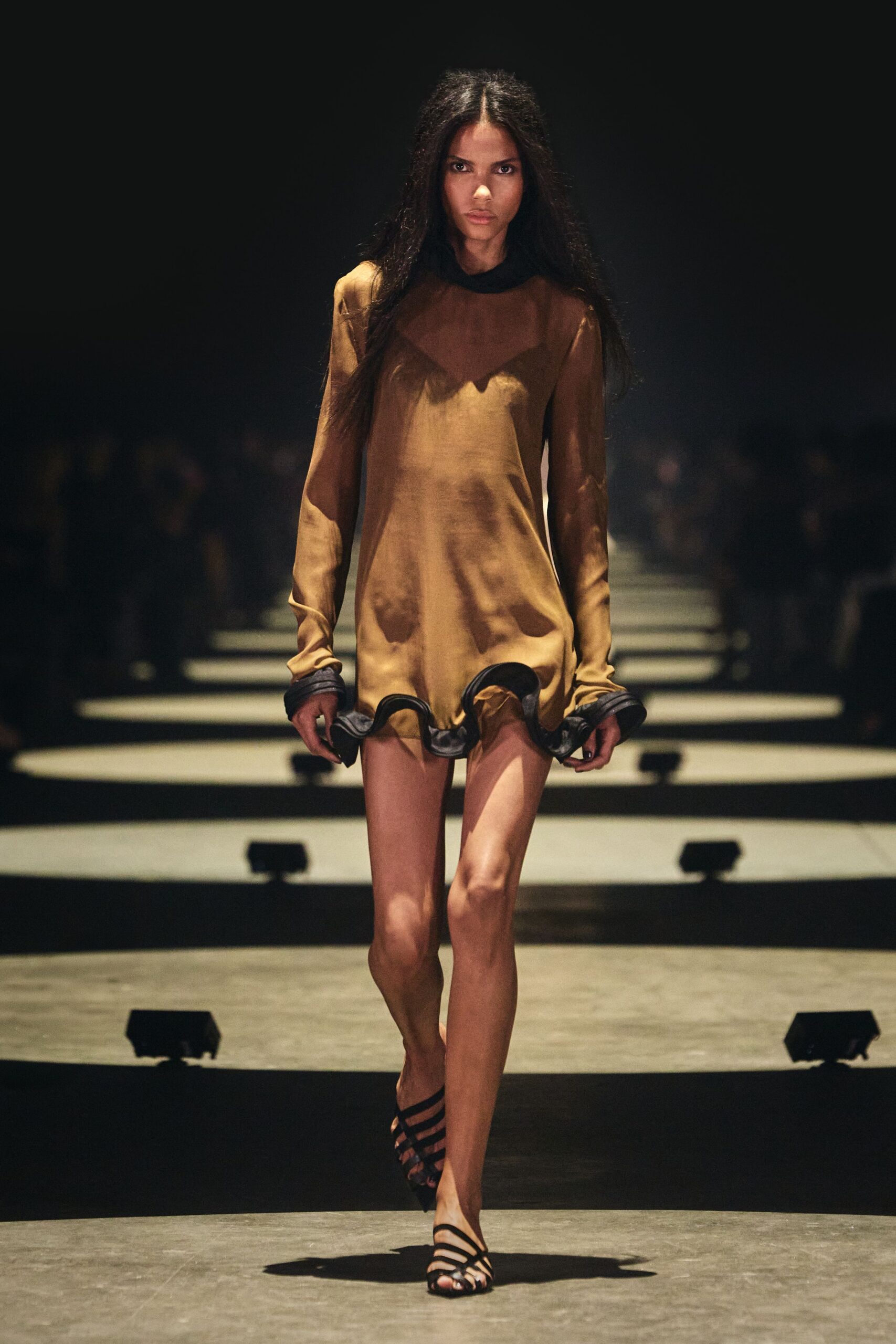 Bronze satin mini dress with sheer yoke and curled black ruffle hem. Black strappy sandals, bands slicing diagonally across the foot. For slipping out of a hotel bar just before the singer's last set.
