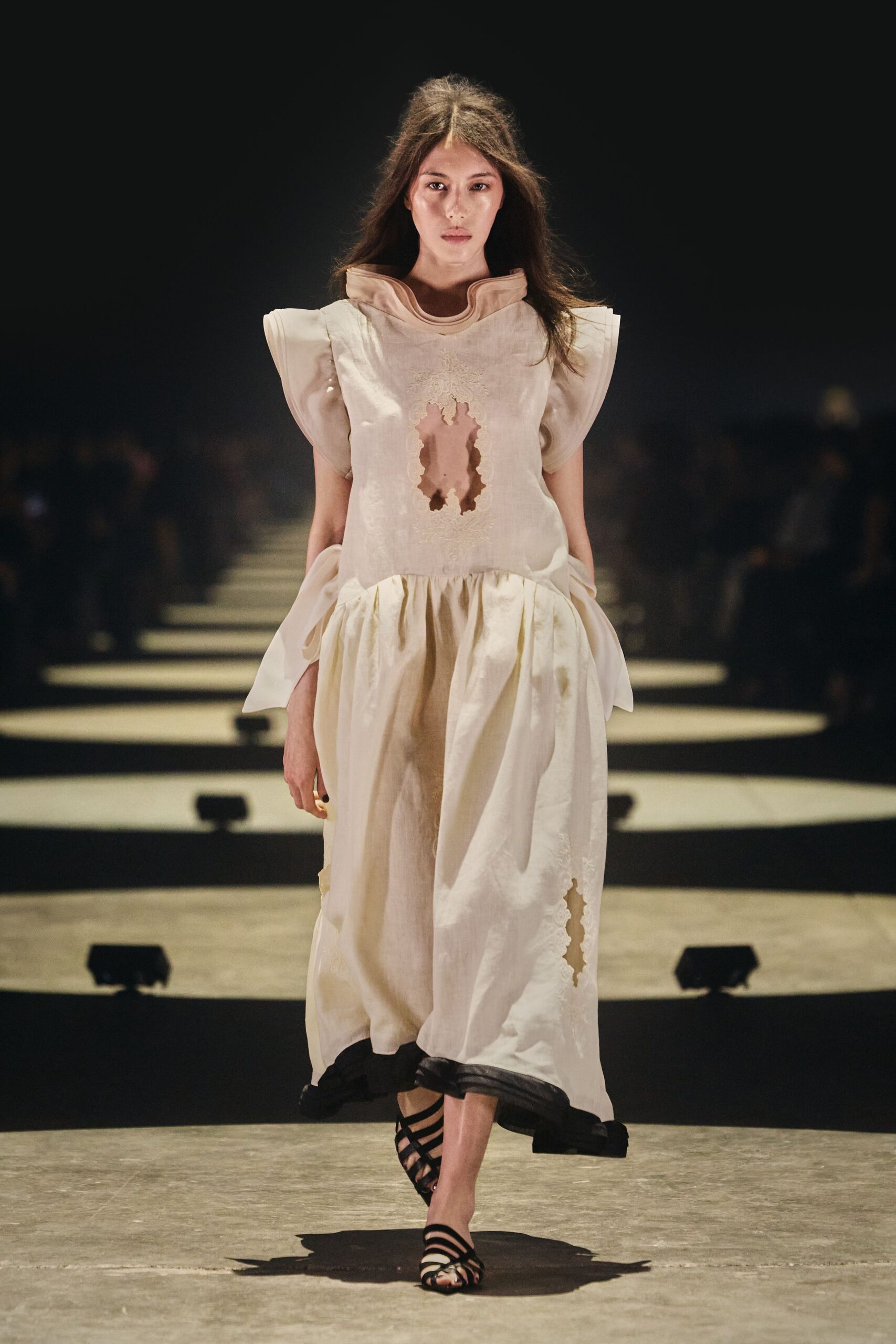 Ivory linen dress with winged sleeves, lace-rimmed holes cut through bodice and skirt. Black strappy sandals with narrow bands crossing the toes. Walking across a museum atrium after someone opened all the windows.