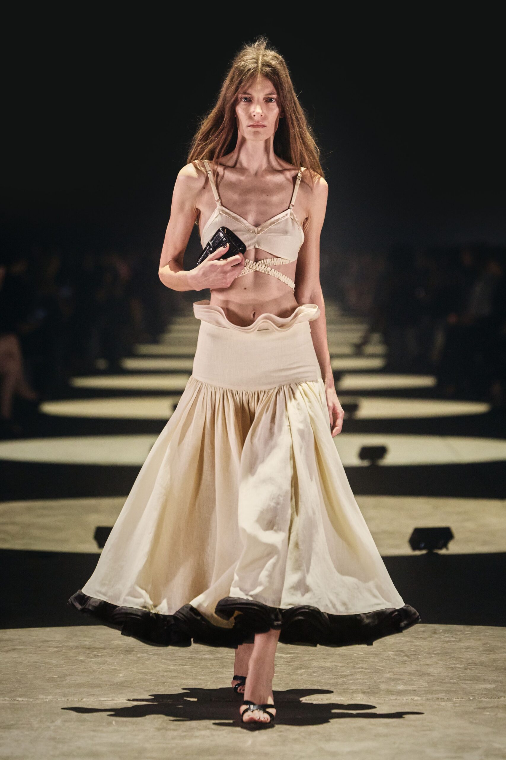 Ivory satin bralette with a ruched band crossing bare ribs. Small black crocodile-embossed clutch gripped flat in one hand. Low-slung ivory skirt, waistband curled into a soft standing ruffle. Black thong sandals with a tiny glint at the toe. For crossing a hotel lobby at dawn with last night still on.