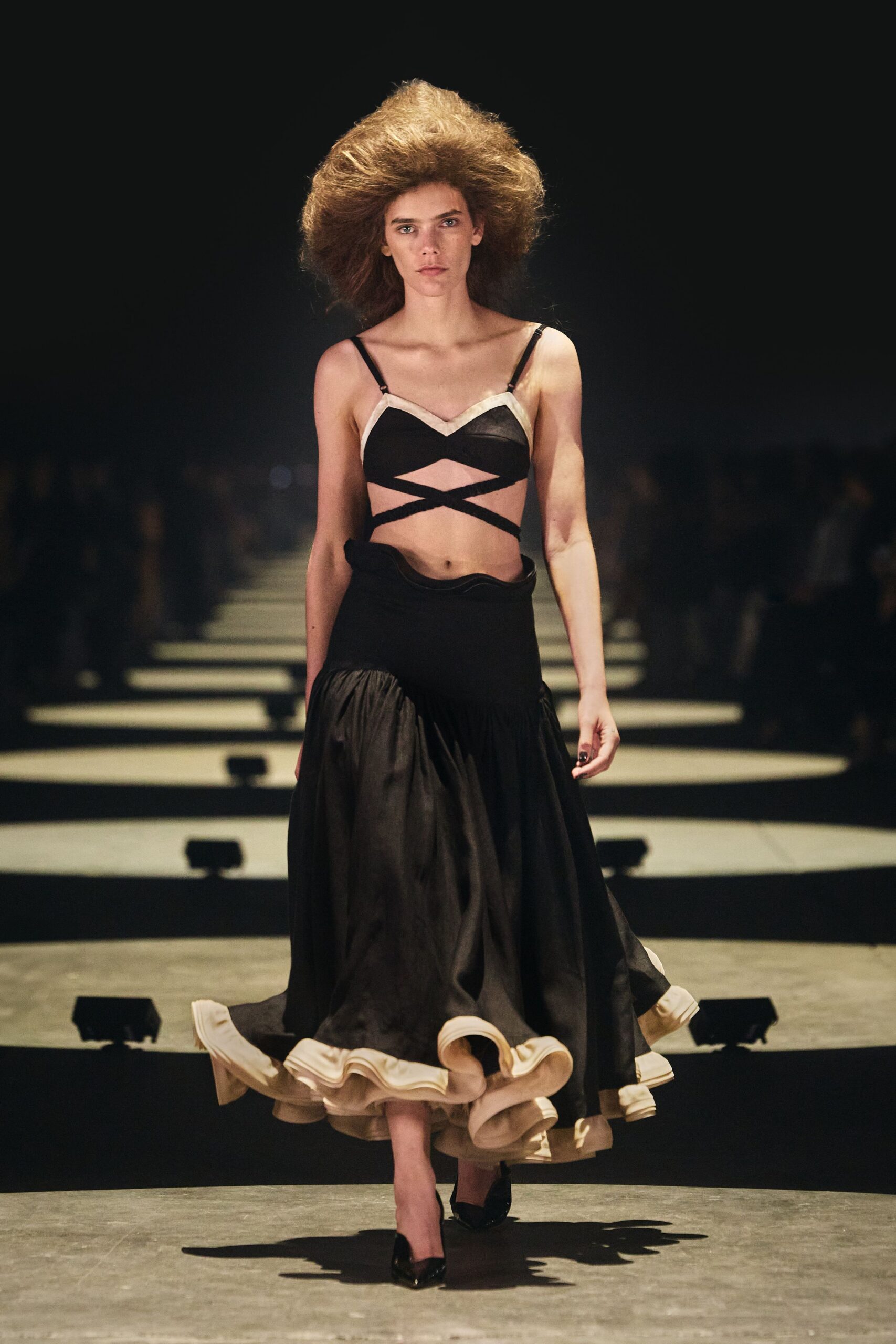 Black satin bra top with ivory binding and crossed bands underbust. Black full skirt dropped from a wavy hip seam, cream flounce curling outward. Black patent pumps. For crossing a hotel ballroom after midnight with one strap already slipping.