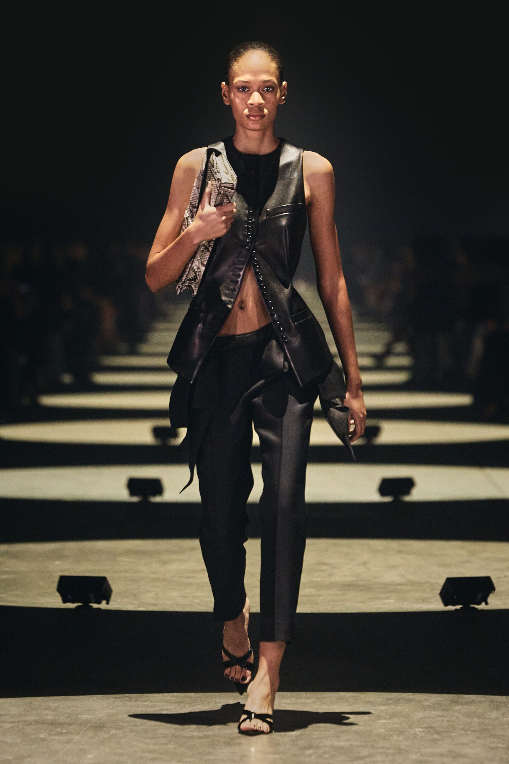 Black leather vest with hook-and-eye closure and flared hip panels. Slim black trousers with a razor crease and low rise. Python shoulder bag tucked tight under one arm. Black patent thong sandals with narrow crossed straps. For slipping into a hotel bar after midnight with nowhere to put your jacket.