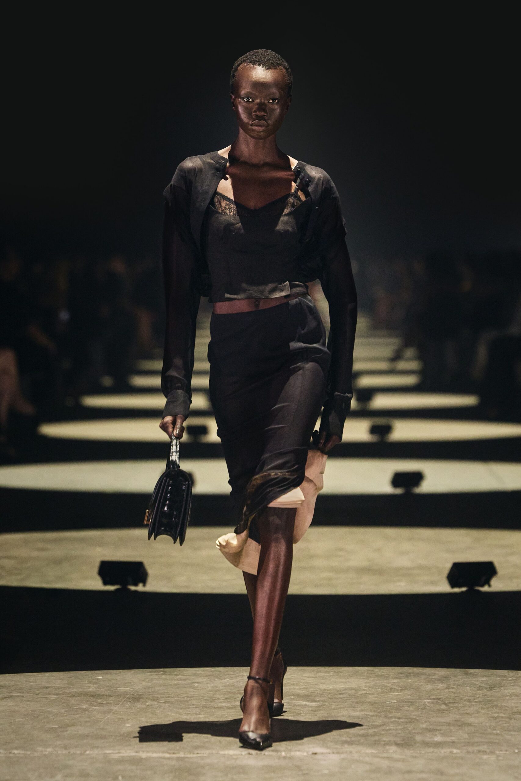 Black fine-knit cardigan hanging open on tiny shoulder buttons. Black satin camisole with scalloped lace peeking at the neckline. Bias black skirt split by a pale underlayer at the hem. Boxy black top-handle bag with accordion gussets. Black ankle-strap pumps, sharply pointed. Whatever you wear to return a key before daylight.