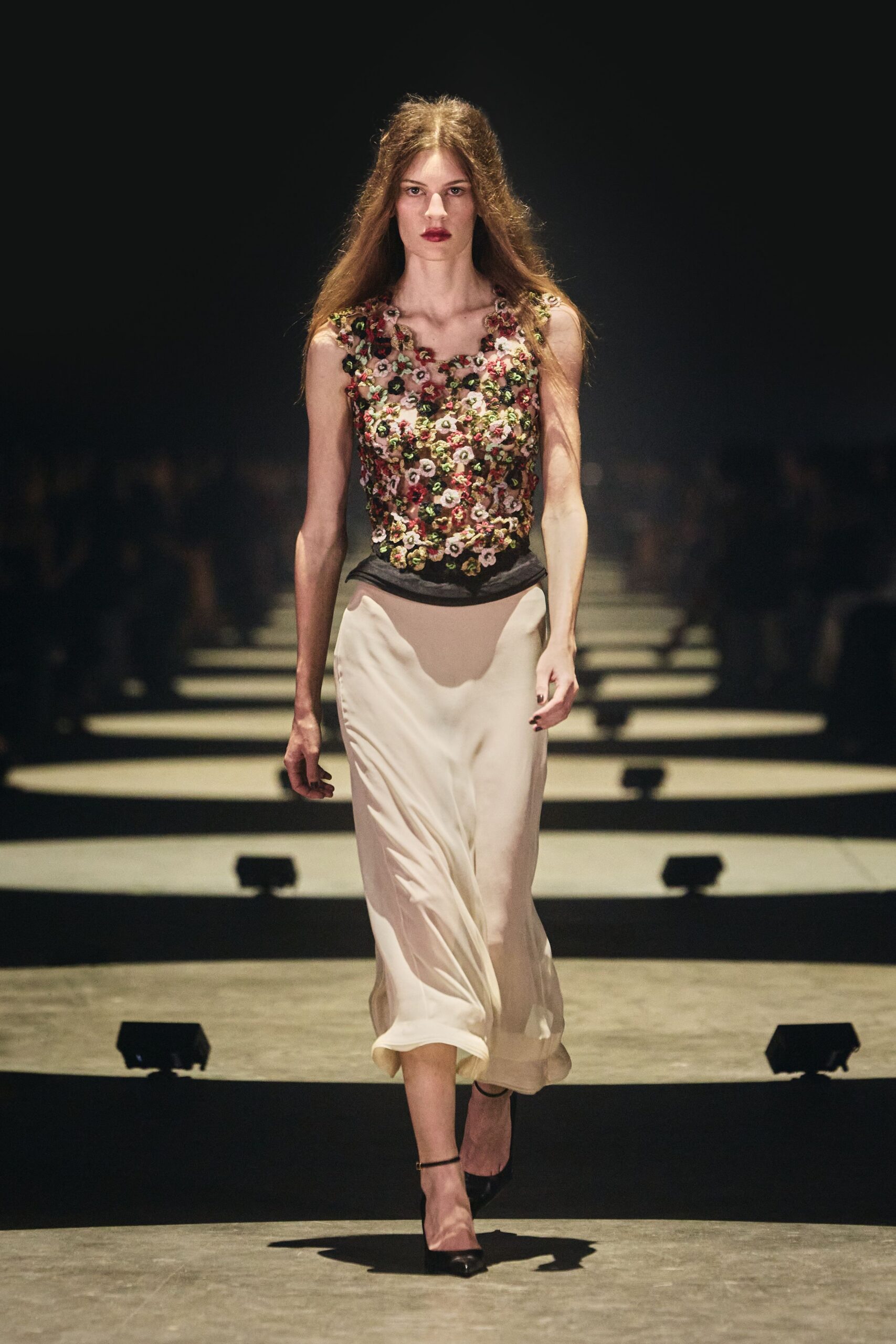Sleeveless mesh top thick with pink and oxblood rosette embroidery. Ivory bias-cut slip skirt, black yoke dipping low at the hip. Black patent ankle-strap pumps. Crossing a hotel ballroom after the flowers have started to drop.