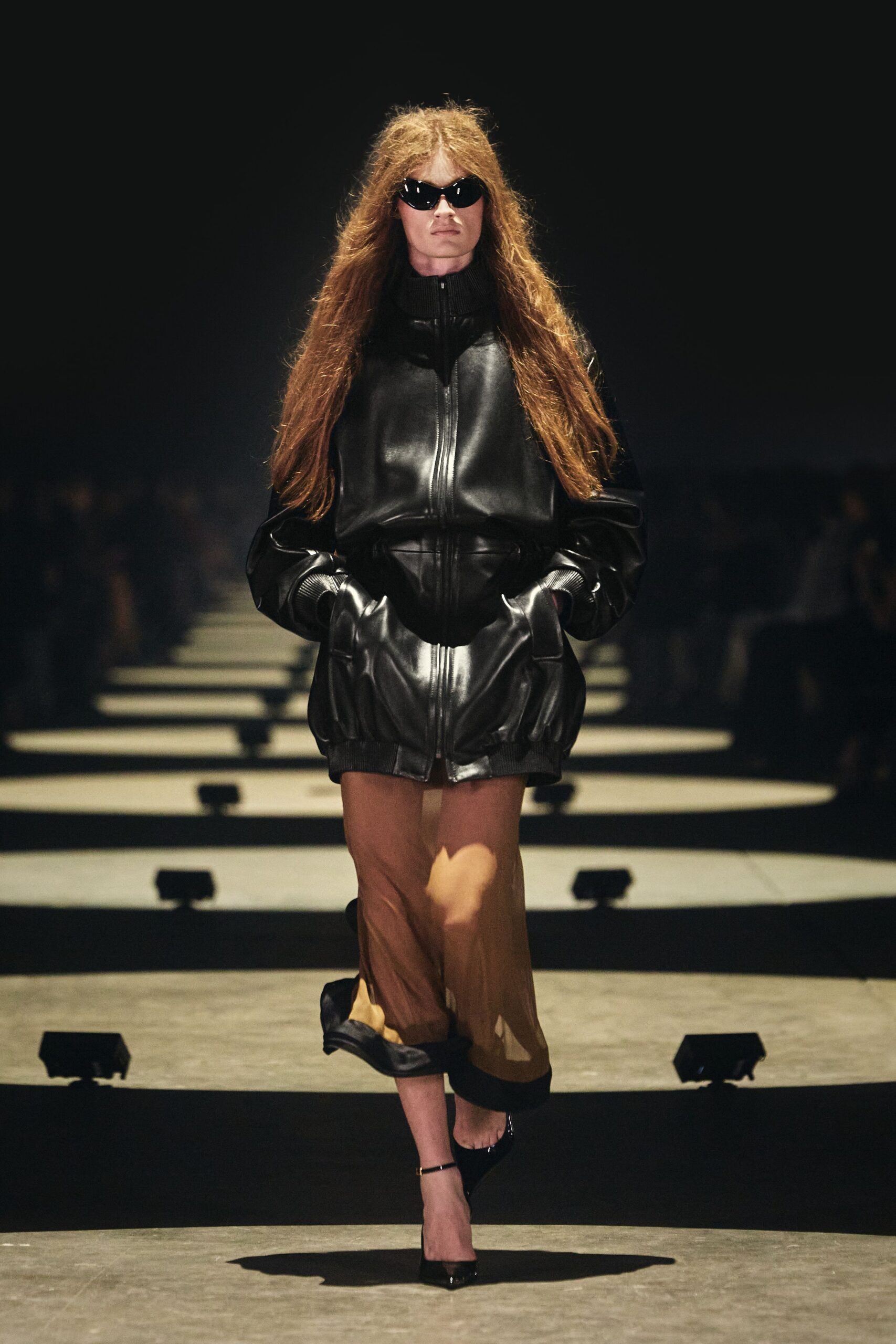 Black wraparound sunglasses with sharp upswept corners. Black leather bomber jacket, doubled zip front and ribbed funnel collar. Sheer tan tube skirt, pooled folds caught under a black satin hem. Black patent pumps with a slim gold ankle strap. Walking into a basement screening after rain, hair still full of weather.