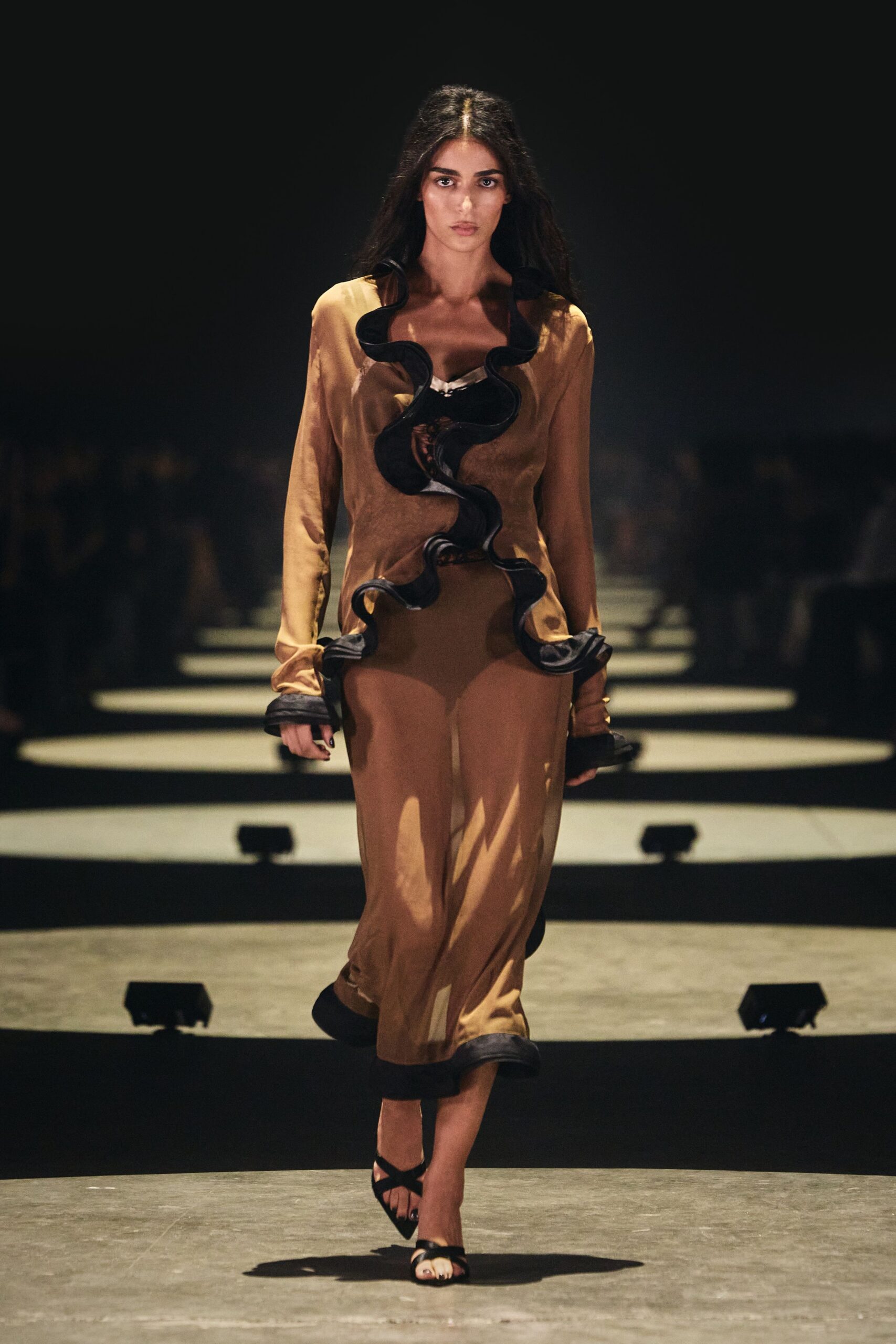 Bronze satin column dress, split by oversized black ruffles curling to the hem. Black strappy heels with a pale toe peeking through. Crossing a hotel lobby after midnight with the flowers already wilting.
