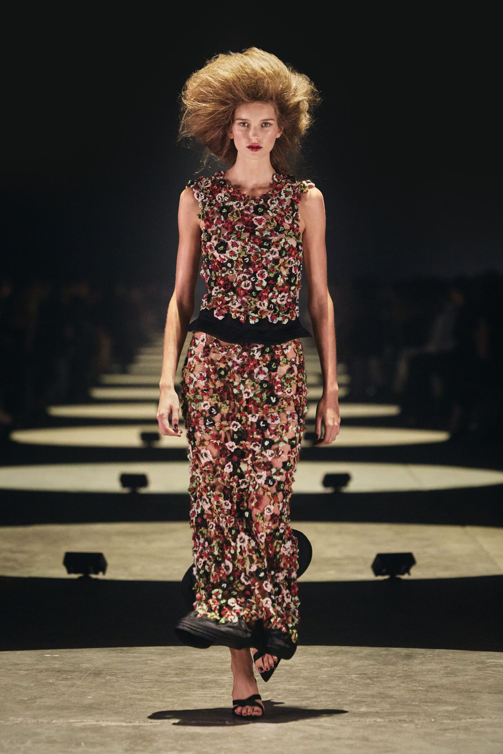 Sleeveless floral appliqué shell, tiny rosettes packed into a raised surface. Matching sheer mesh column, blossoms scattered over nude ground with black godets. Black thong sandals. Walking across a hotel terrace after midnight, petals stuck to the paving.