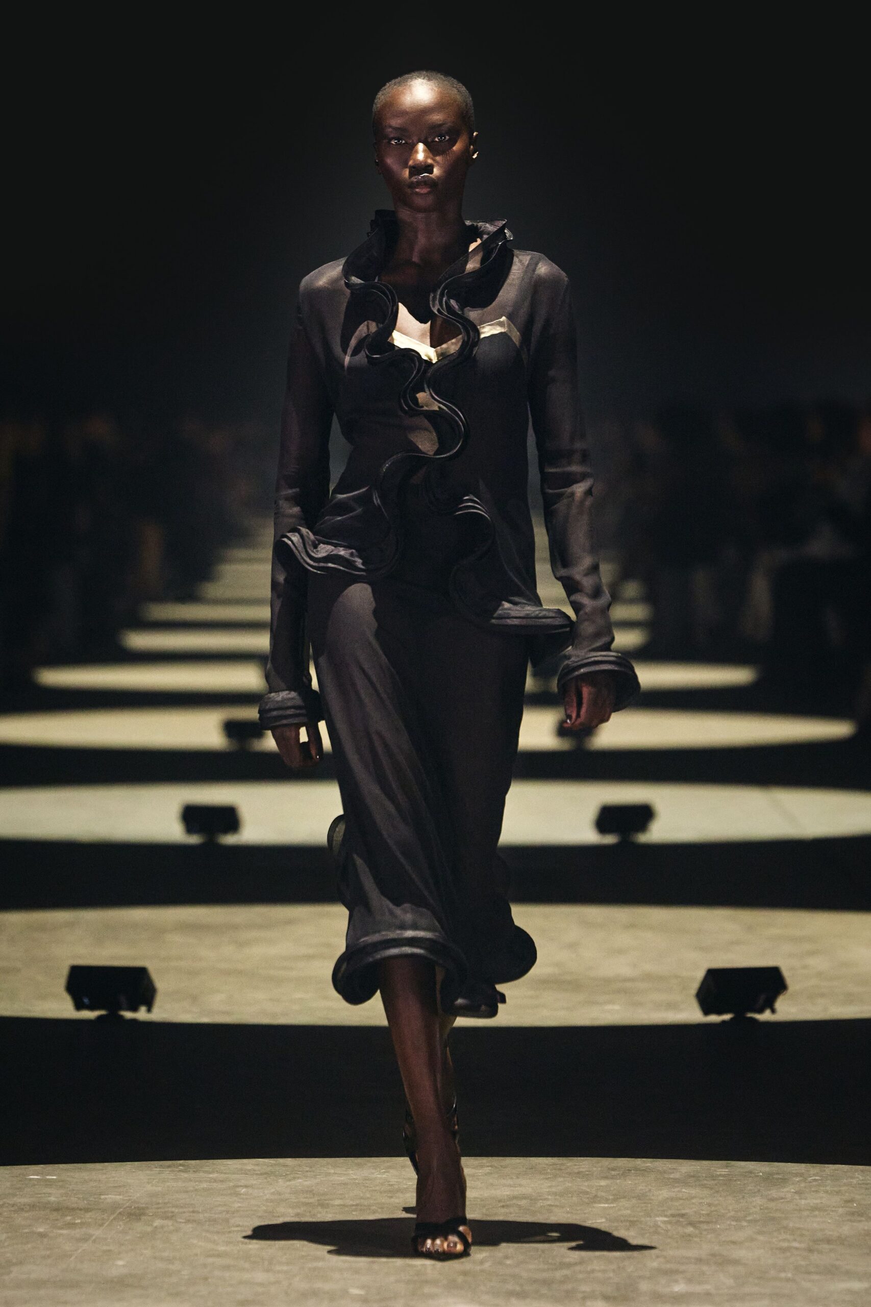 Smoke-black column gown with a towering collar and wired ruffles spiraling off-center. Black open-toe heels, barely there under the long hem. Crossing a hotel lobby at midnight after the string quartet packed up.