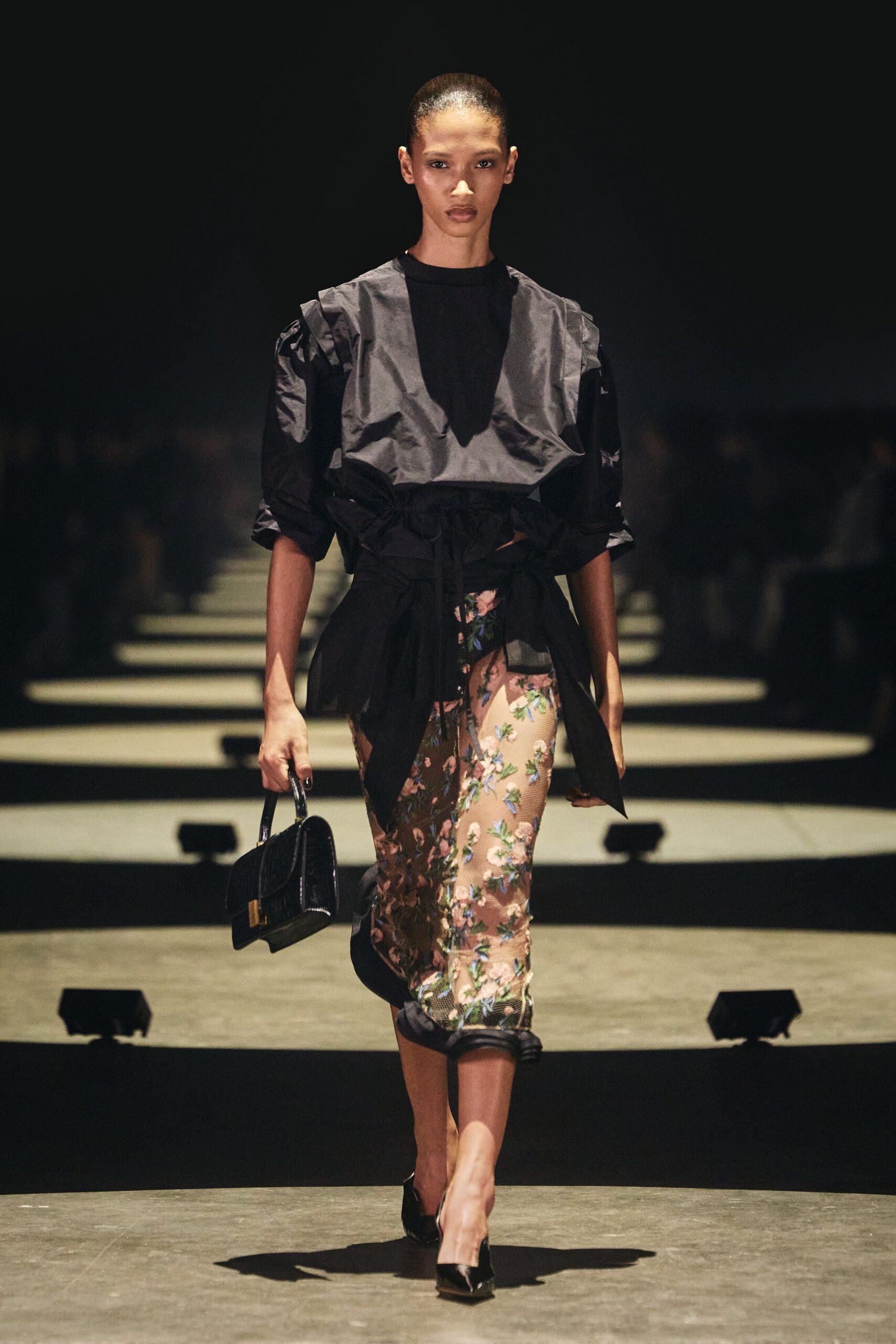 Black taffeta top with a deep V bib and puffed sleeves. Paperbag waist cinched with long self ties. Nude mesh pencil skirt scattered with pink and blue blossoms. Black patent top-handle bag with a gold clasp. Black patent pointed pumps. Walking into a hotel bar while the wedding upstairs is still going.