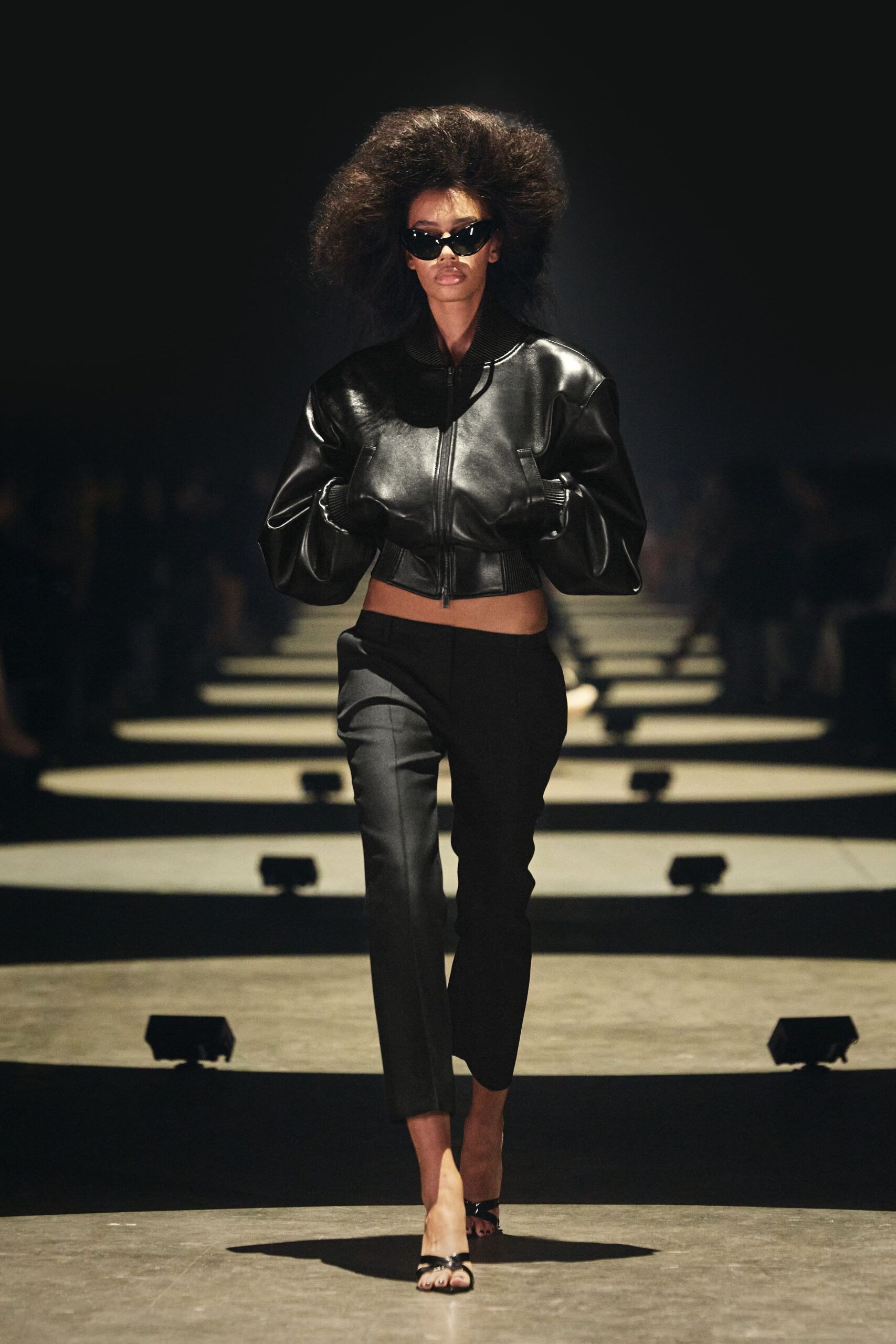 Narrow black cat-eye frames with a sharp glossy tilt. Cropped black leather bomber, ribbed collar and hem cinching hard. Black cigarette trousers with a pressed crease and cropped ankle. Barely there black patent toe-ring sandals. Walking through a shuttered arcade before the cleaners unlock the gates.