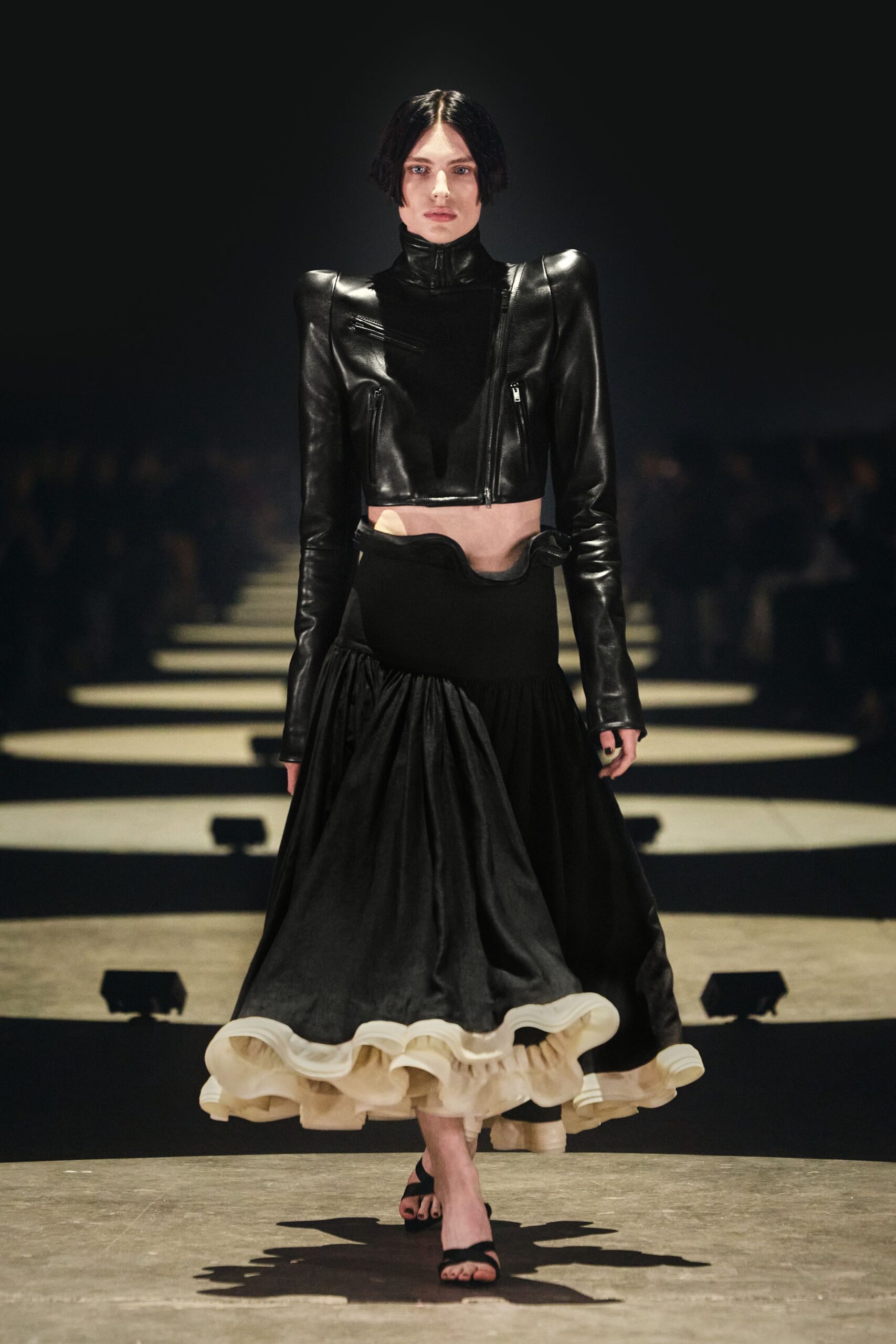 Cropped black leather jacket with peaked shoulders and three exposed zips. Black drop-waist skirt, waistband cut into a scooped wave at center. Cream organza flounce curling out beneath the black skirt. Bare black two-strap sandals. Crossing a marble foyer after midnight, carrying the argument with you.