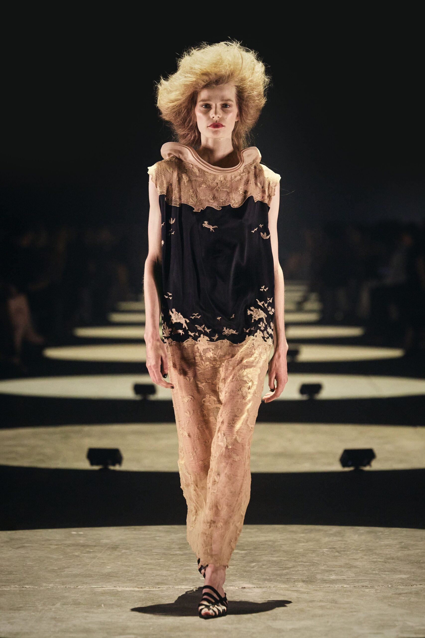 Sleeveless column dress with a nude rolled neckline and ragged lace yoke. Black silk center panel scattered with tiny ivory birds and hares. Black caged sandals, narrow straps banding the toes. Crossing a hotel lobby at dawn with last night's eyeliner still on.
