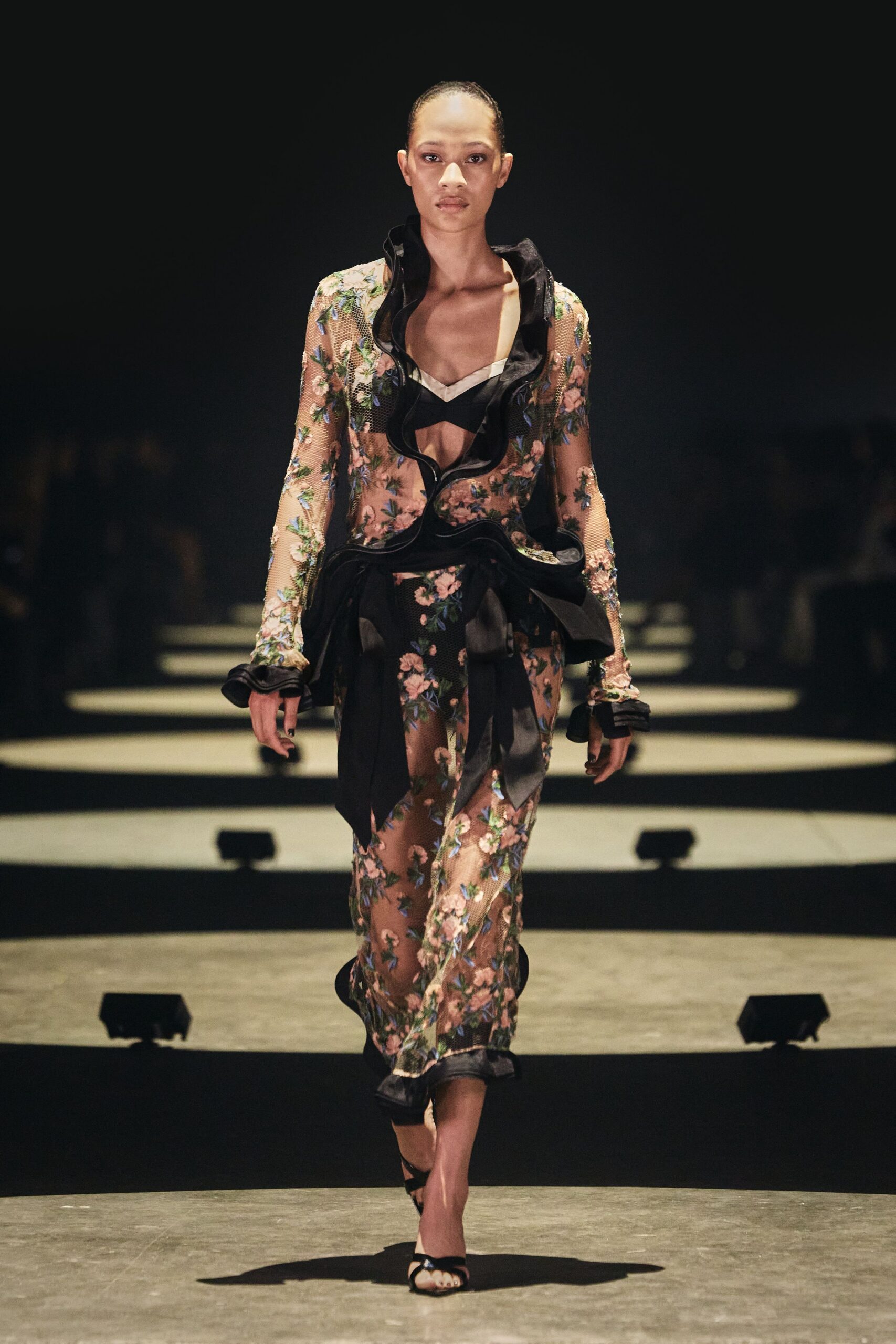 Sheer nude mesh column densely embroidered with pink blossoms and blue leaves. Black satin ruffles whip around the deep V and hips. Black bra top with a pale ivory notch at the neckline. Black satin ankle-strap sandals. Walking through a winter conservatory after dark, petals stuck to the damp floor.