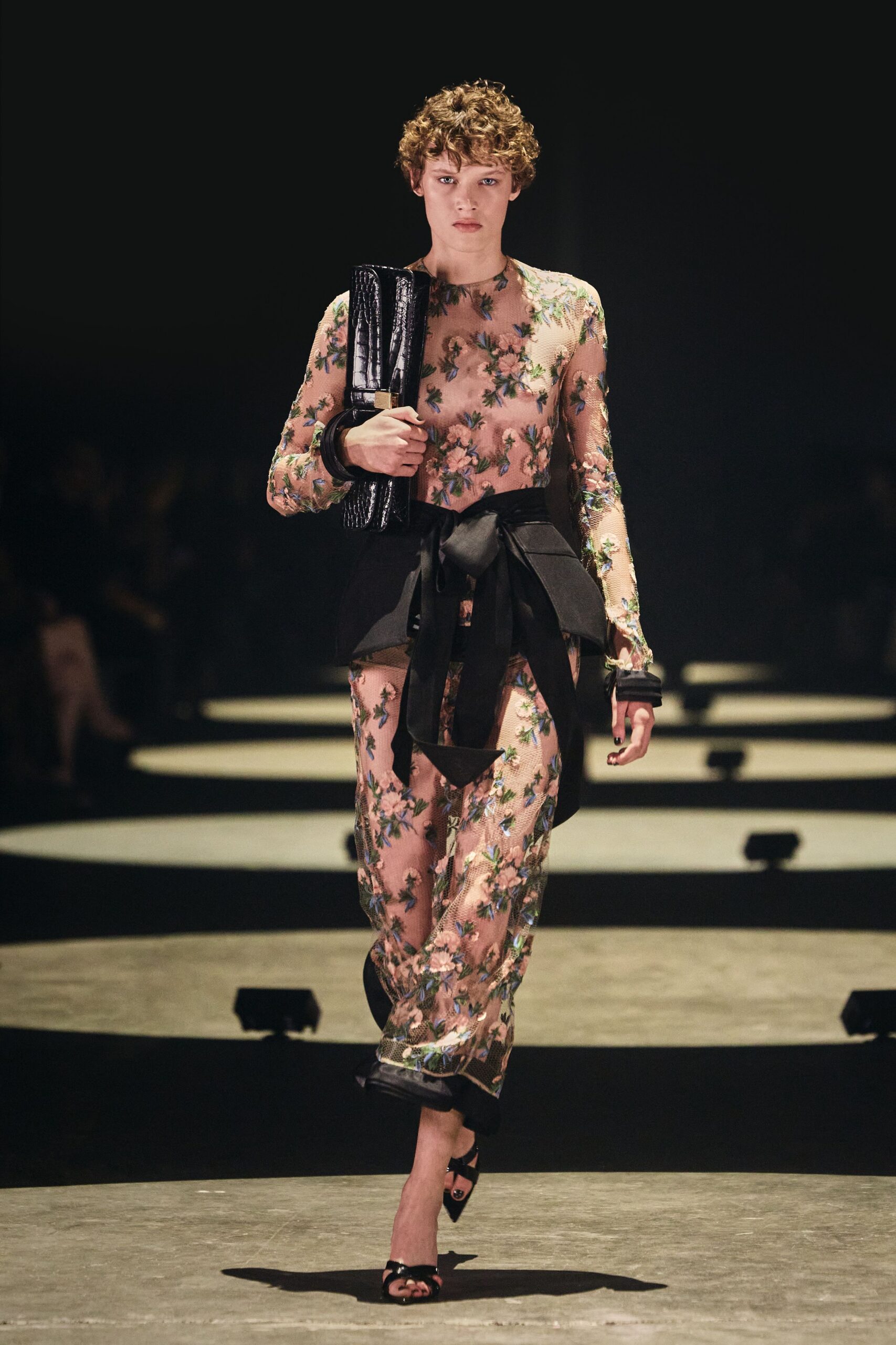 Sheer nude mesh jumpsuit scattered with embroidered pink blossom and blue sprigs. Black peplum belt tied low, one long sash dropping past the knee. Glossy black crocodile-embossed clutch tucked upright under the arm. Black patent toe-ring sandals. Crossing a hotel atrium after midnight with somebody else's room key.