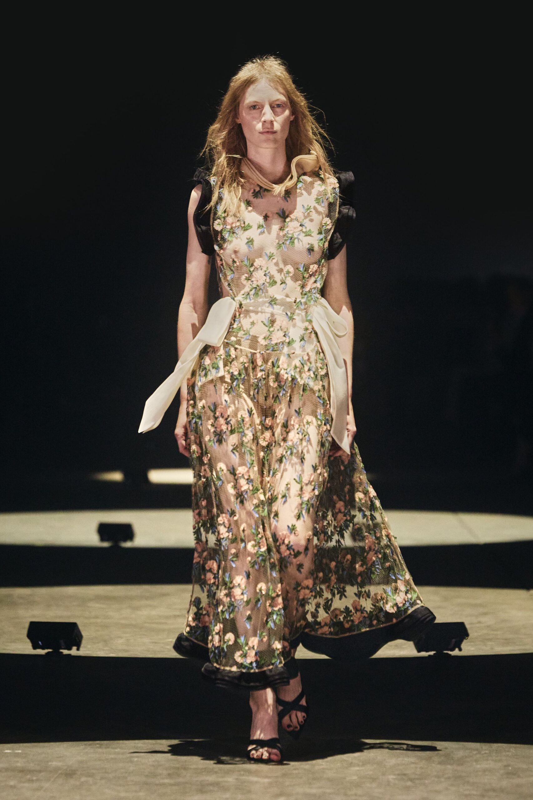 Blush mesh dress densely embroidered with trailing flowers over a pale slip. Cream ties looped at the hips, one tail hanging longer. Black ankle-strap heels. Walking across a hotel lawn after the wedding, carrying your shoes by midnight.
