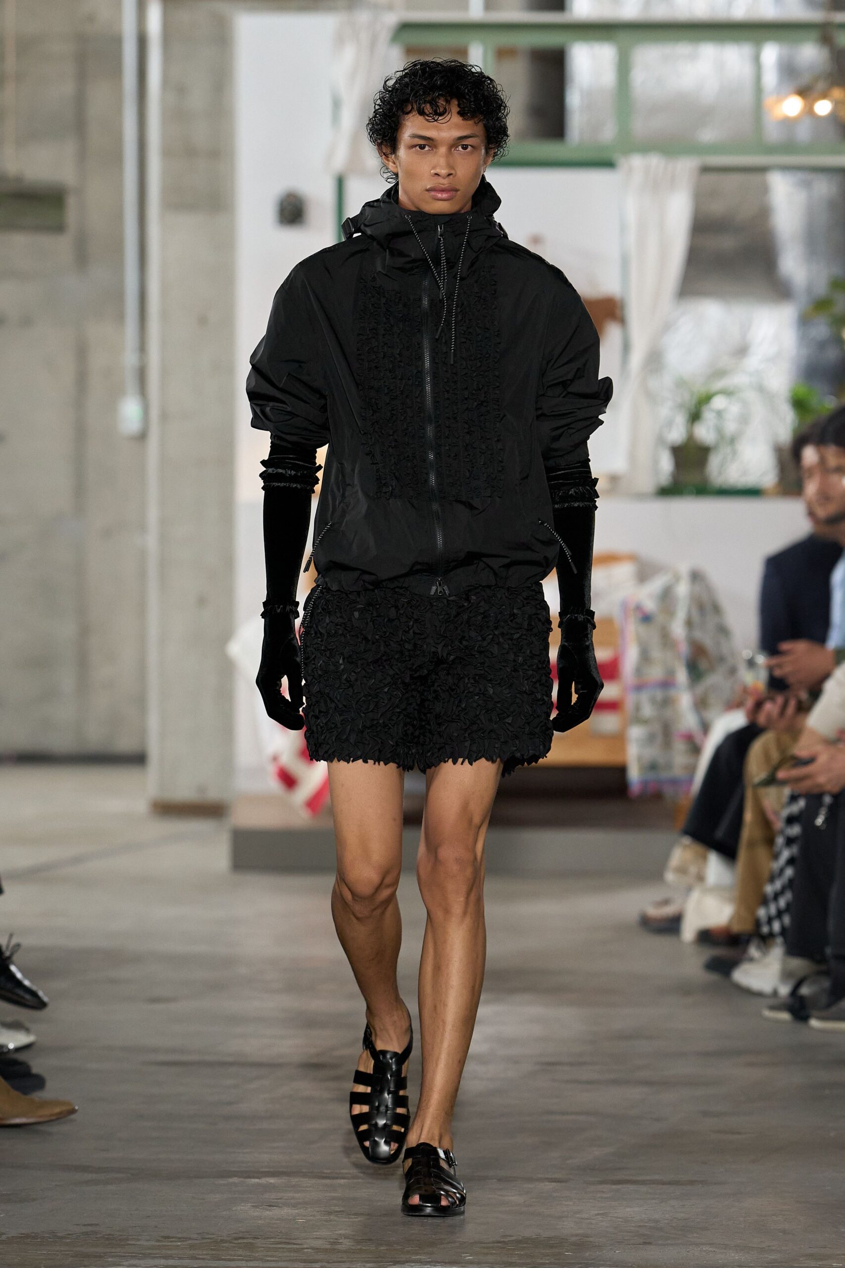 Black shell jacket with a ruffled bib panel and two-way zip. Elbow-length black velvet gloves, softly crushed at the wrists. Black shorts densely covered in curled appliqué, hem sitting uneven. Black caged leather sandals with a slingback strap. Crossing a tiled courtyard after rain, carrying nothing but your room key.
