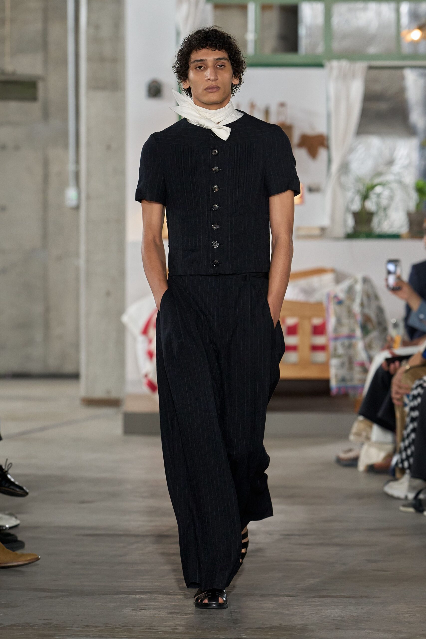 White voile neck scarf knotted low, one pointed end standing up. Black pinstriped top with fine pintucks and a column of dark buttons. Loose black pinstriped trousers breaking softly over the foot. Black leather cage sandals. For crossing a tiled veranda before lunch is called in.