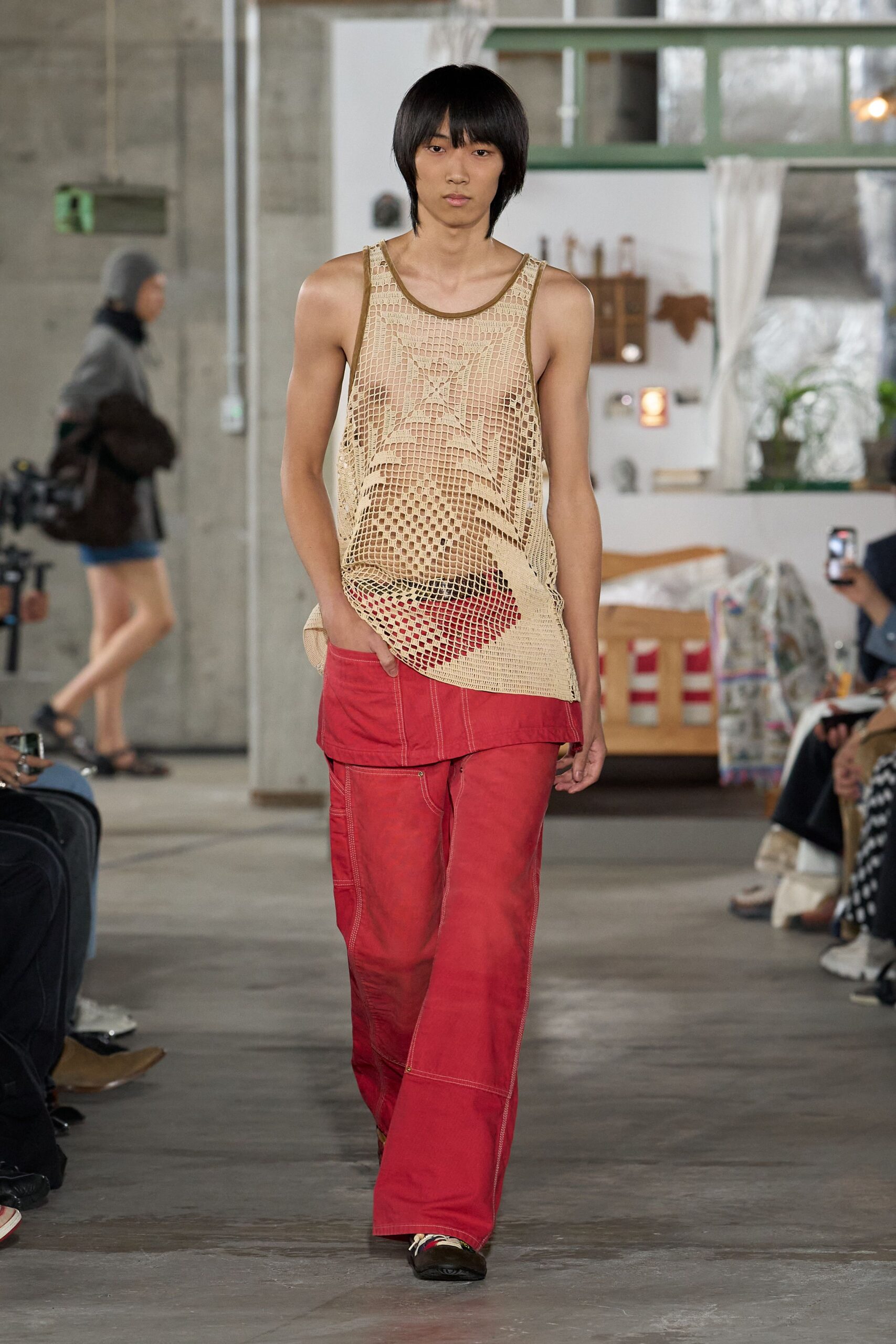 Loose beige openwork tank, crochet squares sagging over the torso. Red underlayer with a curved flap hem peeking below. Faded red workwear trousers with pale topstitching and panel seams. Black slip-ons with white laces dragged loose. Walking back from the beach with salt drying on your shoulders.