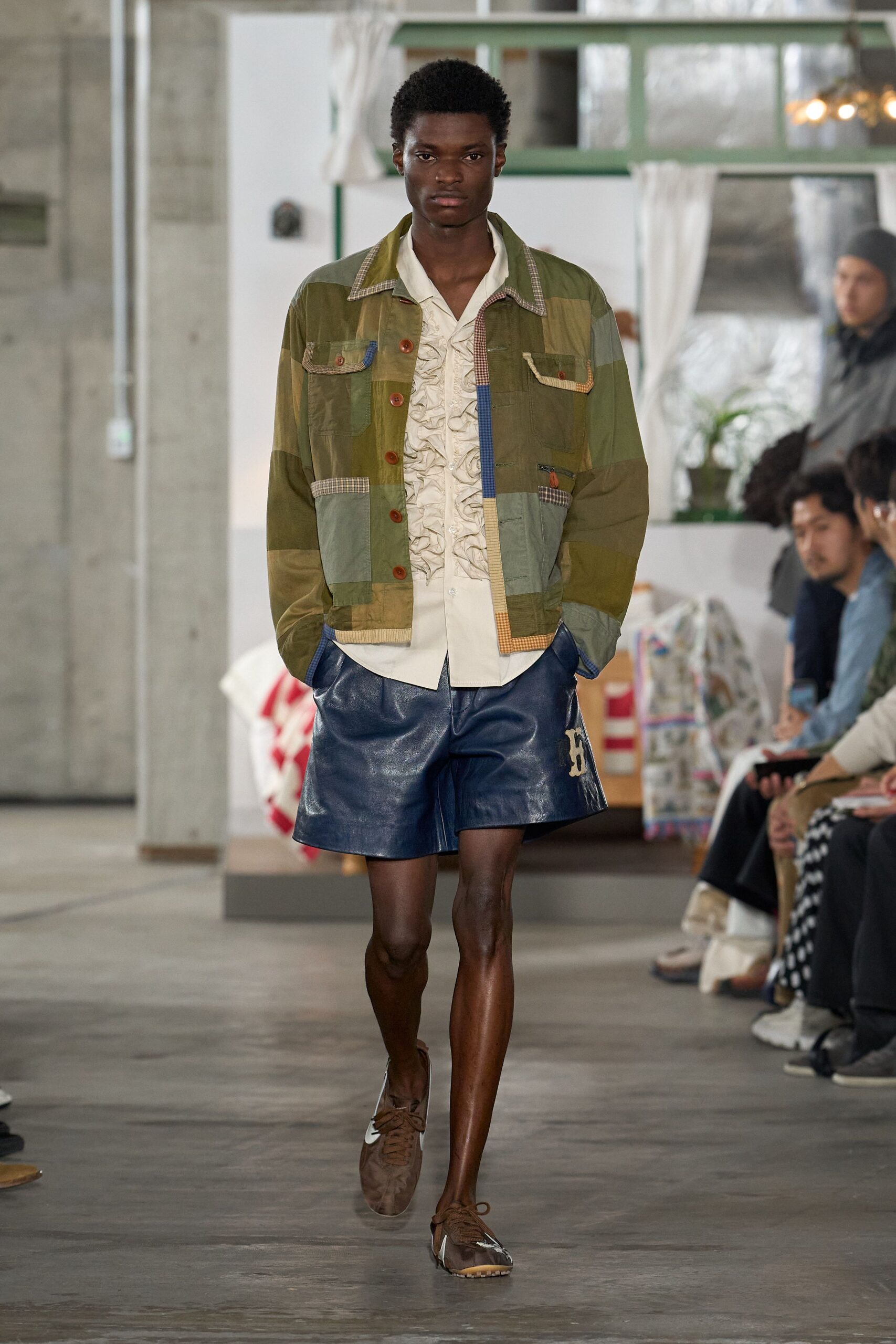 Olive patchwork jacket with mismatched checks edging the flap pockets. Cream shirt front packed with narrow ruffles. Cobalt leather shorts with a pale appliqued patch at one leg. Brown low-top sneakers with translucent side panels. Walking back from the lake house store with cards and bread.