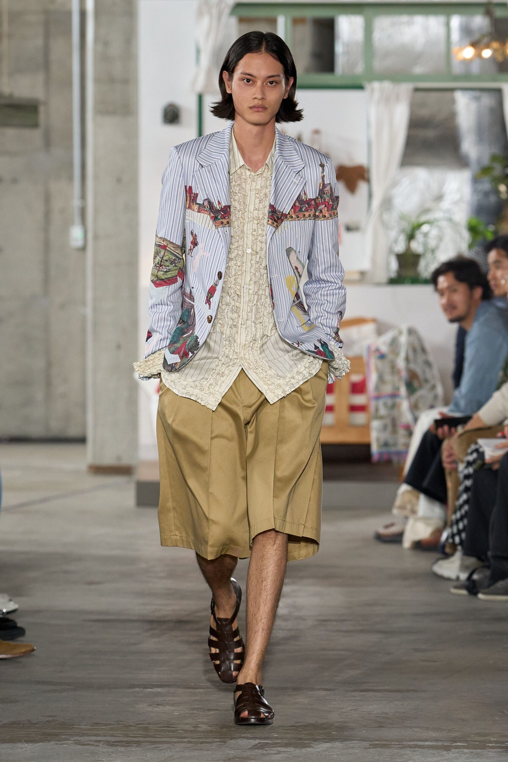 Powder-blue pinstripe blazer printed with village scenes across the chest. Cream striped shirt fronted with dense ruffles, cuffs spilling past the sleeves. Khaki pleated shorts cut wide and long below the knee. Dark brown leather fisherman sandals. Sunday house tour, someone has left the porch door open to the heat.