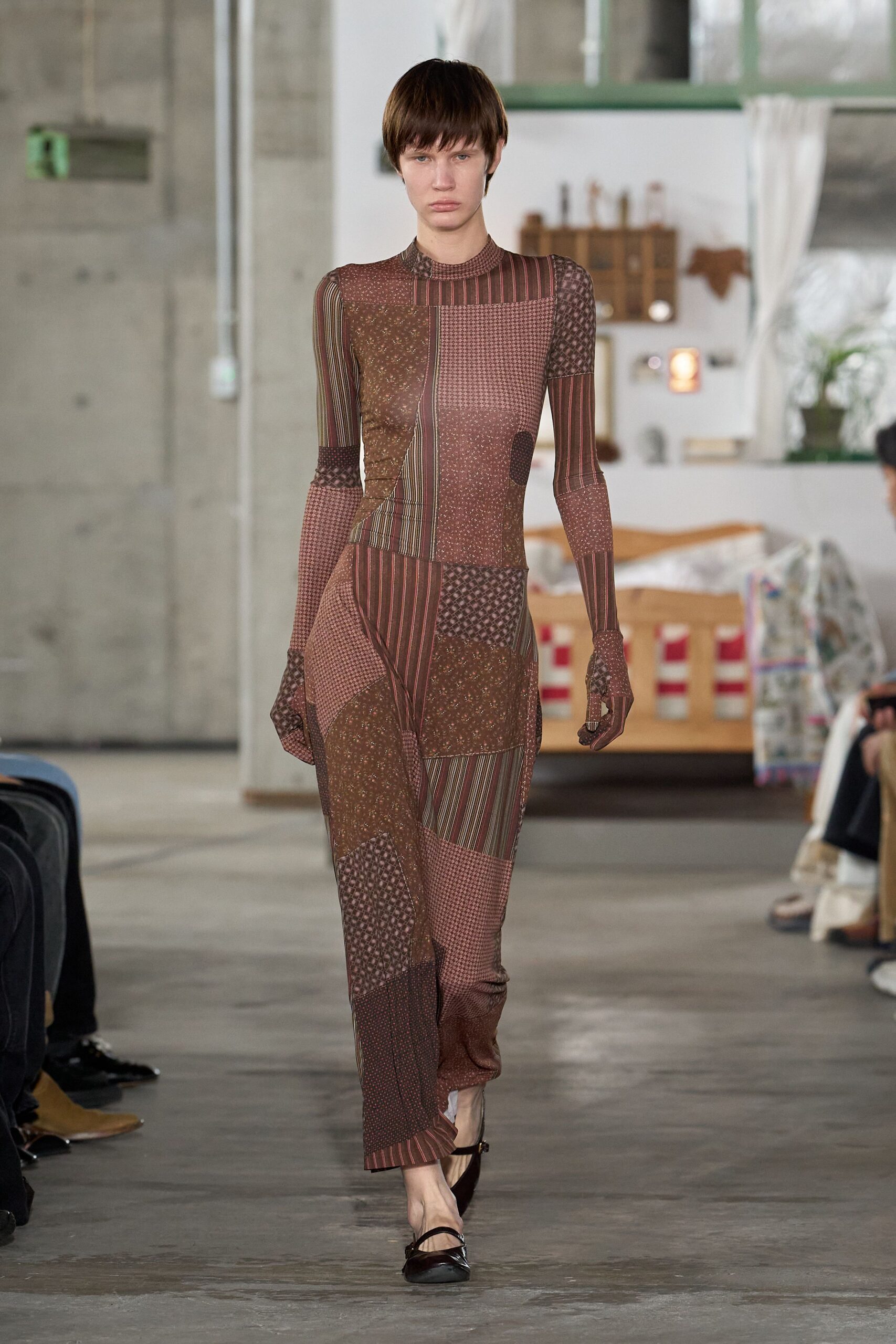 Second-skin patchwork knit dress in cocoa and mauve, pieced from mixed tiny geometrics and pinstripes. Built-in glove sleeves, thumbholes pulling the print over the hands. Dark patent Mary Jane flats with a narrow strap. Walking across cold concrete after sunrise, still wearing last night's patterned jersey.