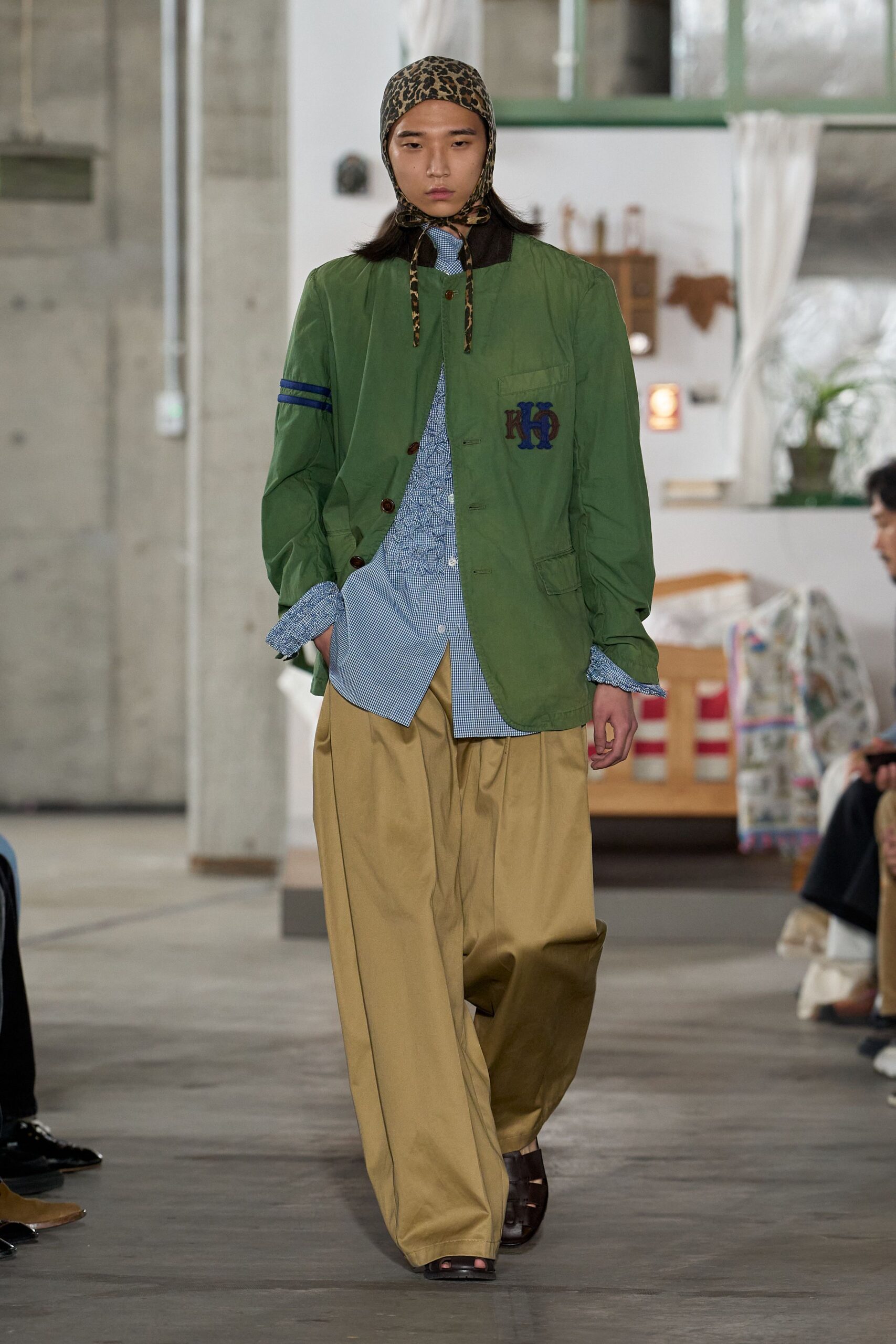 Leopard scarf tied under the chin, long ends hanging loose. Moss cotton jacket with striped arm bands and a monogram patch. Blue micro-check shirt, ruffled placket peeking through the opening. Camel wide-leg trousers with deep front pleats. Dark brown fisherman sandals. Walking into a rural post office just before lunch closes.