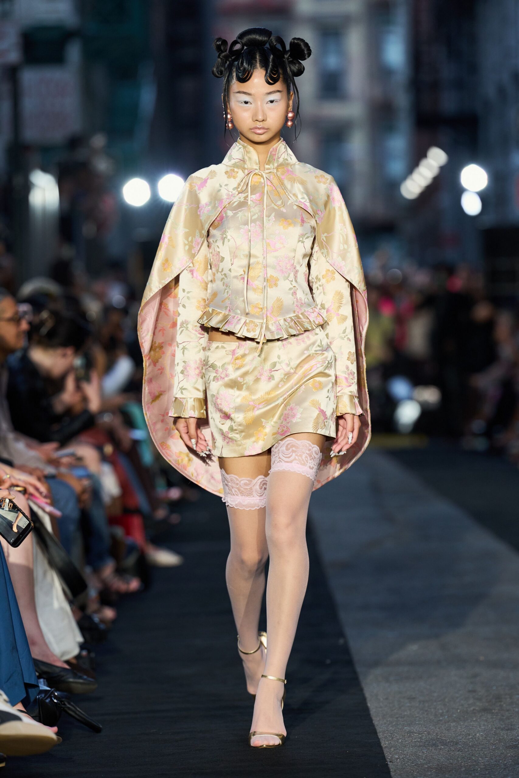 Champagne floral satin cape blouse, frog-tied collar and peplum ruffle. Matching satin mini, cut close with a slight side ripple. Powder-pink lace bands gripping sheer nude thighs. Barely there gold ankle-strap sandals. Pink orb drop earrings. Crossing Chinatown after rain, takeaway box warming your hands.