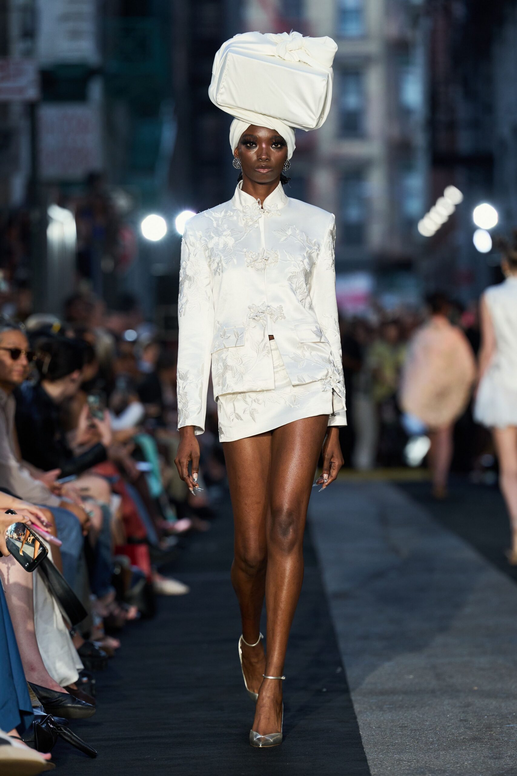 Oversized white satin box balanced over a wrapped headscarf. Ivory brocade jacket with frog closures and a stand collar. Matching wrap mini, one corner kicked slightly upward. Round crystal cluster studs. Silver slingback pumps with clear pointed vamps. Crossing city blocks after the wedding, still carrying the cake box.