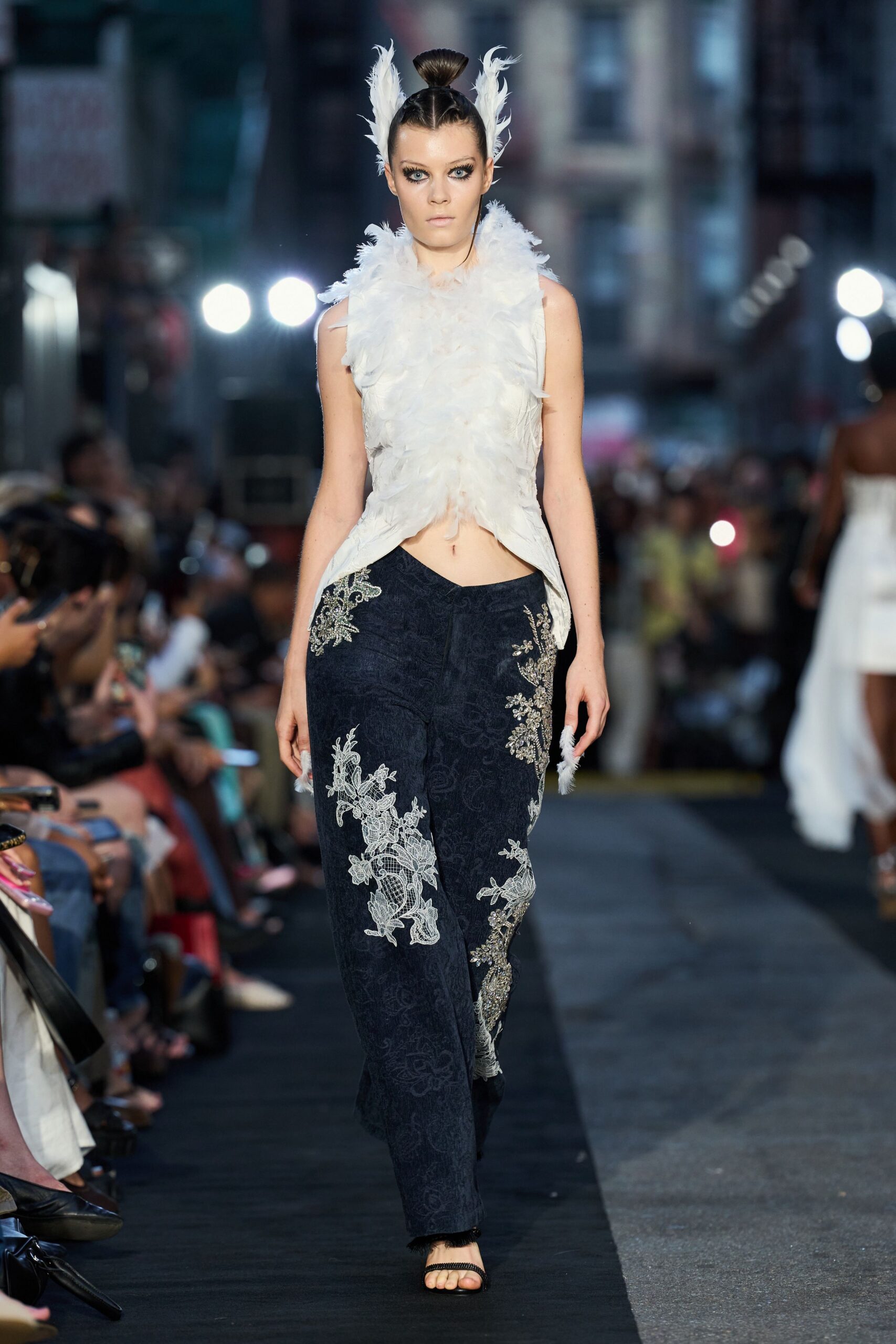 Ivory feather-covered shell, cut away at the waist with pointed front hems. Two upright white feather sprays framing a tight topknot. Inky jacquard trousers with silver lace appliqué climbing one thigh. Black thong sandals with narrow glittered straps. Crossing a hotel forecourt after midnight with confetti still in your hair.