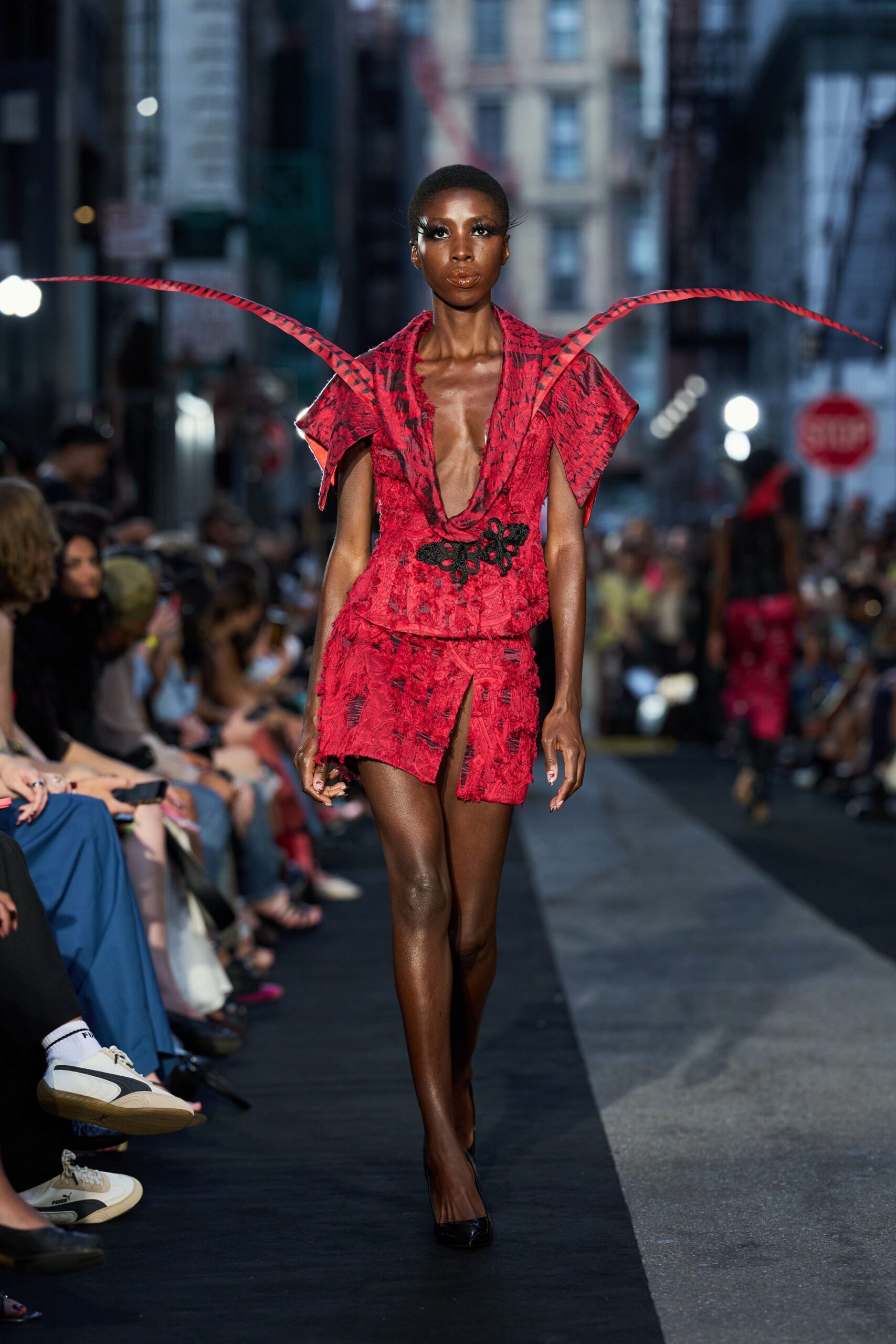 Crimson brocade top with a plunging cowl and winged shoulder flanges. Black corded appliqué cinching the waist like a knotted flourish. Matching frayed brocade mini, split high at the thigh. Black patent pumps. Crossing a blocked-off avenue just after the parade has passed.