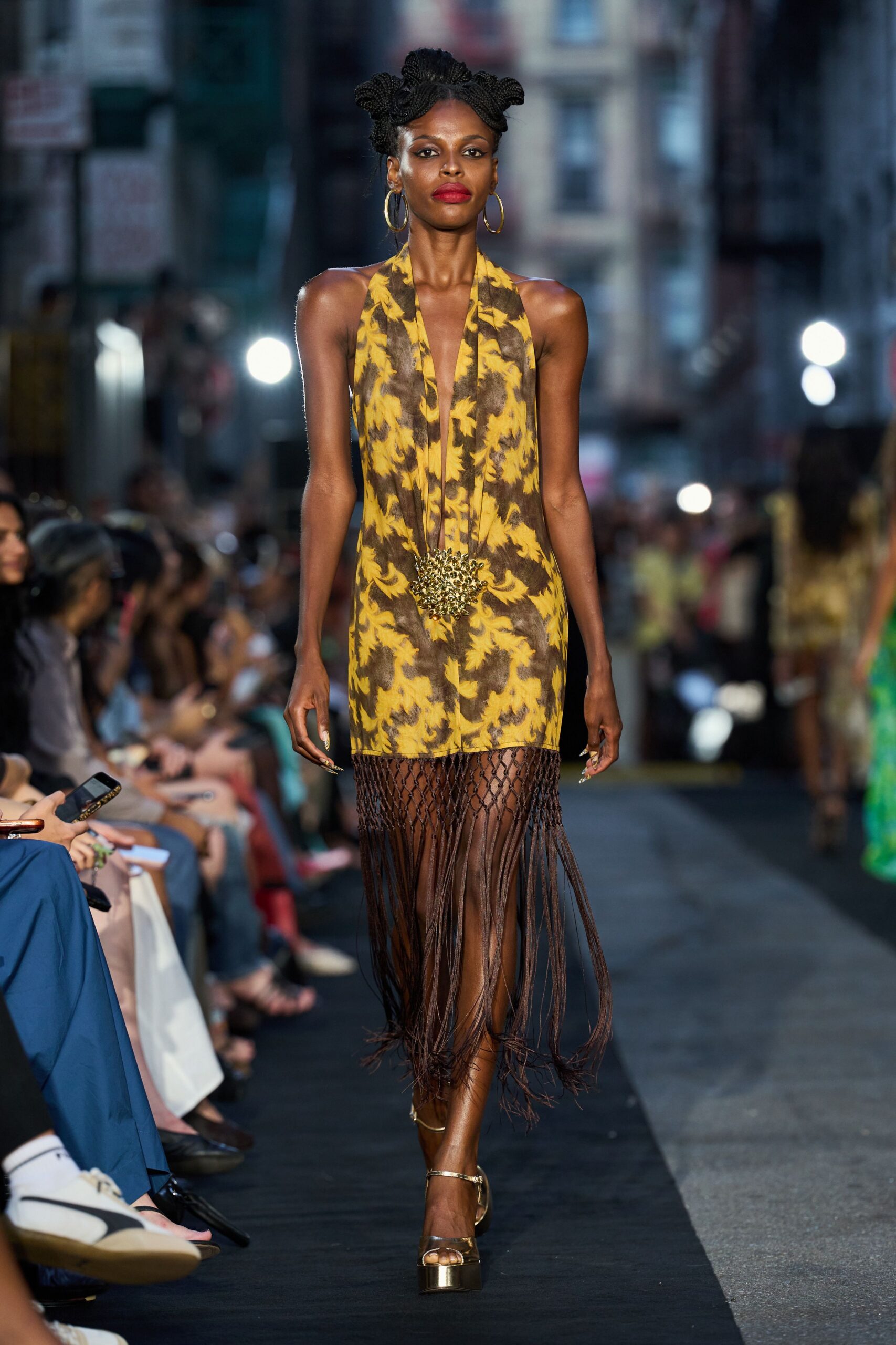 Large gold hoops brushing the jaw. Ochre and umber baroque-print halter dress, plunged to the waist. Spiky gold pendant hanging from a narrow neck cord. Brown knotted net dropping into calf-length rope fringe. Bronze ankle-strap platforms. Walking out of a street party just before the band packs up.