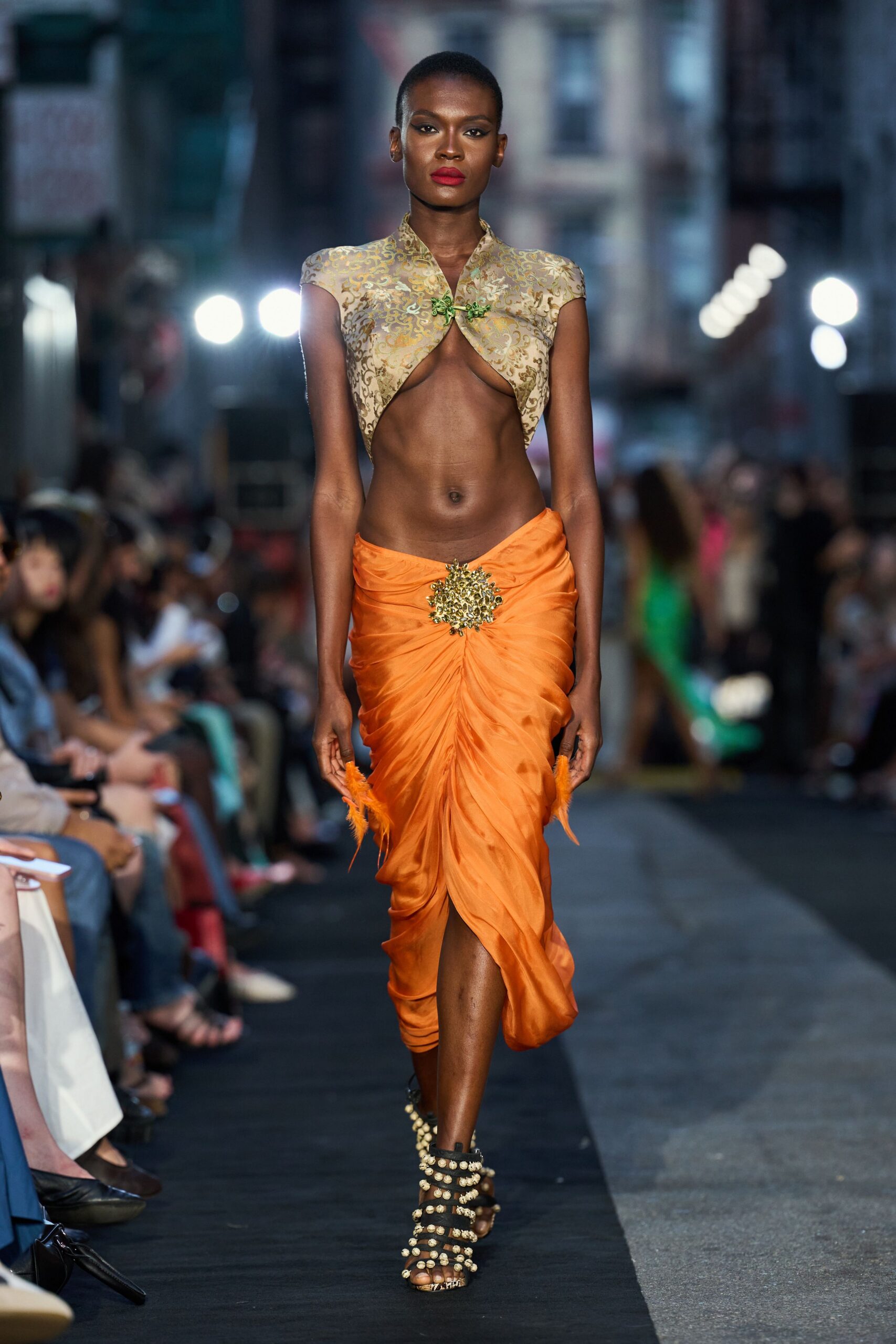 Champagne brocade bolero with cap sleeves and a green frog closure. Tangerine satin sarong skirt, ruched to a jewelled burst at center. Black lace-up sandals studded with rows of shell-like beads. Street festival after sundown, the food stalls still smoking.
