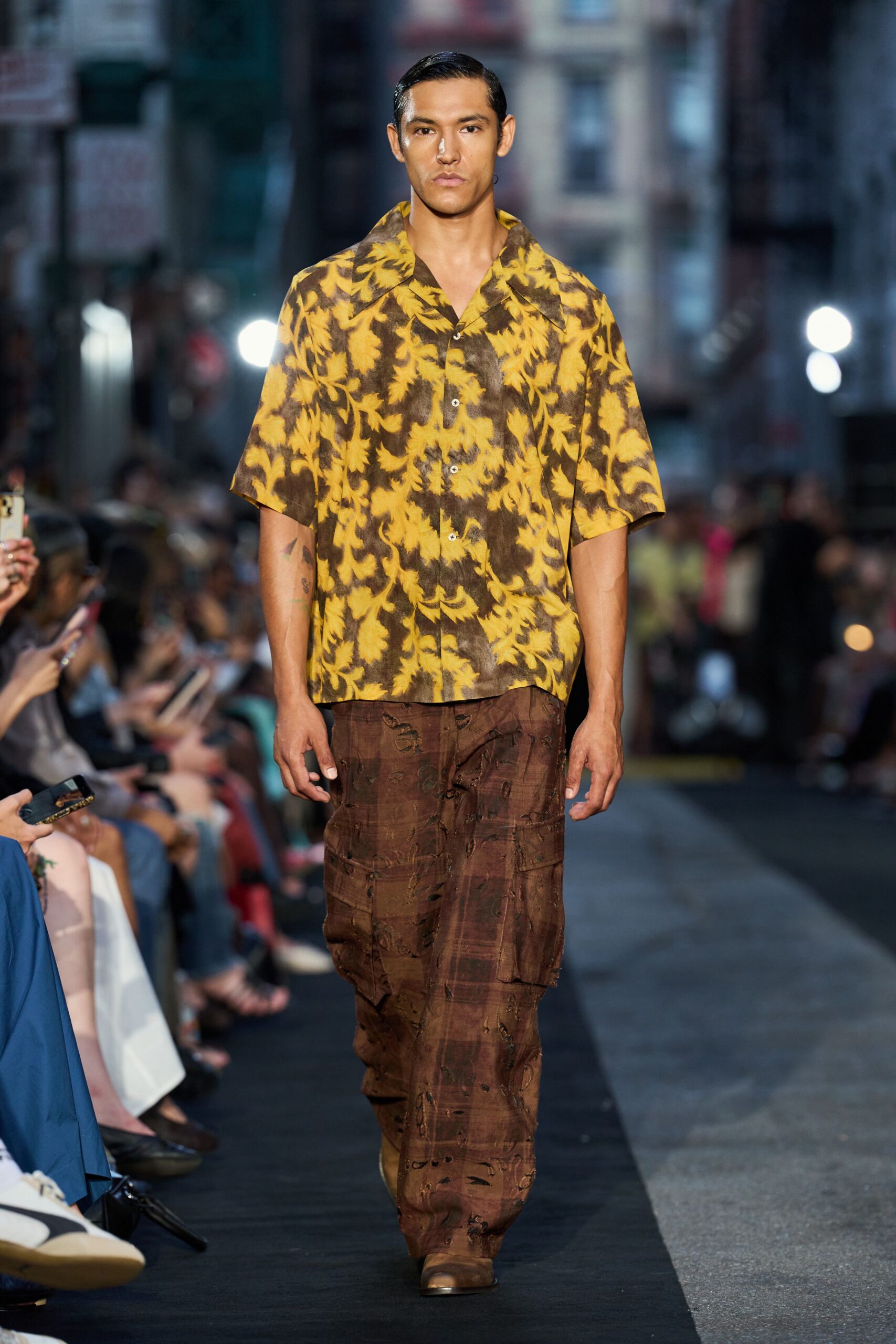 Ochre camp-collar shirt in smoky leaf print, white buttons down the placket. Brown plaid cargo trousers, slashed and laddered across the thigh pockets. Dusty tan boots with blunt square toes. Walking back from a roadside bar after rain, moths still at the neon.