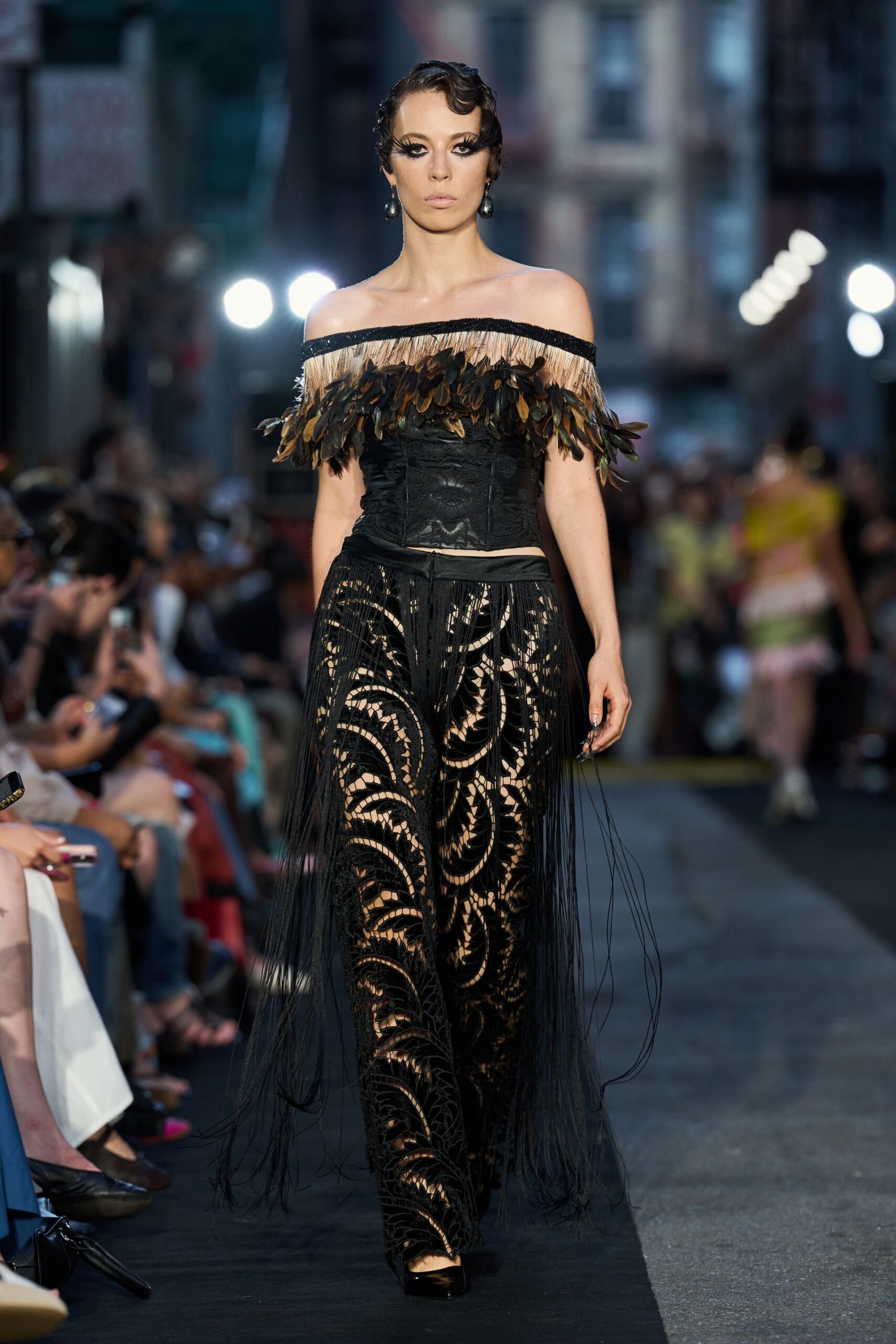 Gunmetal drop earrings with oversized black pearl teardrops. Black corset topped with straw fringe and layered brown-black feathers. Sheer black lace trousers in scrolling cutwork, extra-long fringe from the hips. Black patent pumps, square at the toe. Crossing a harbor square after dark while buskers are packing up.