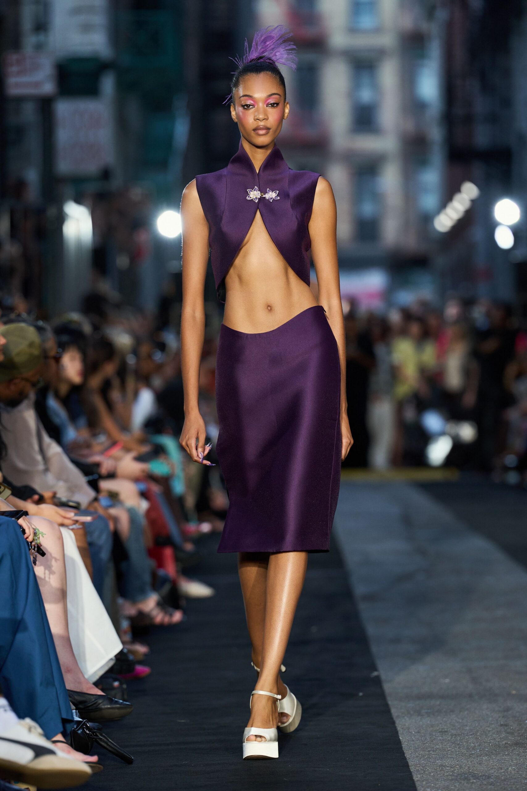 Purple feather spray rising from a tight topknot. Plum satin cropped shell, split to the navel under two orchid clasps. Matching bias-cut midi skirt, waistband dipping low over one hip. White platform sandals with a slim ankle strap. Crossing downtown at dusk after changing out of something much stricter.
