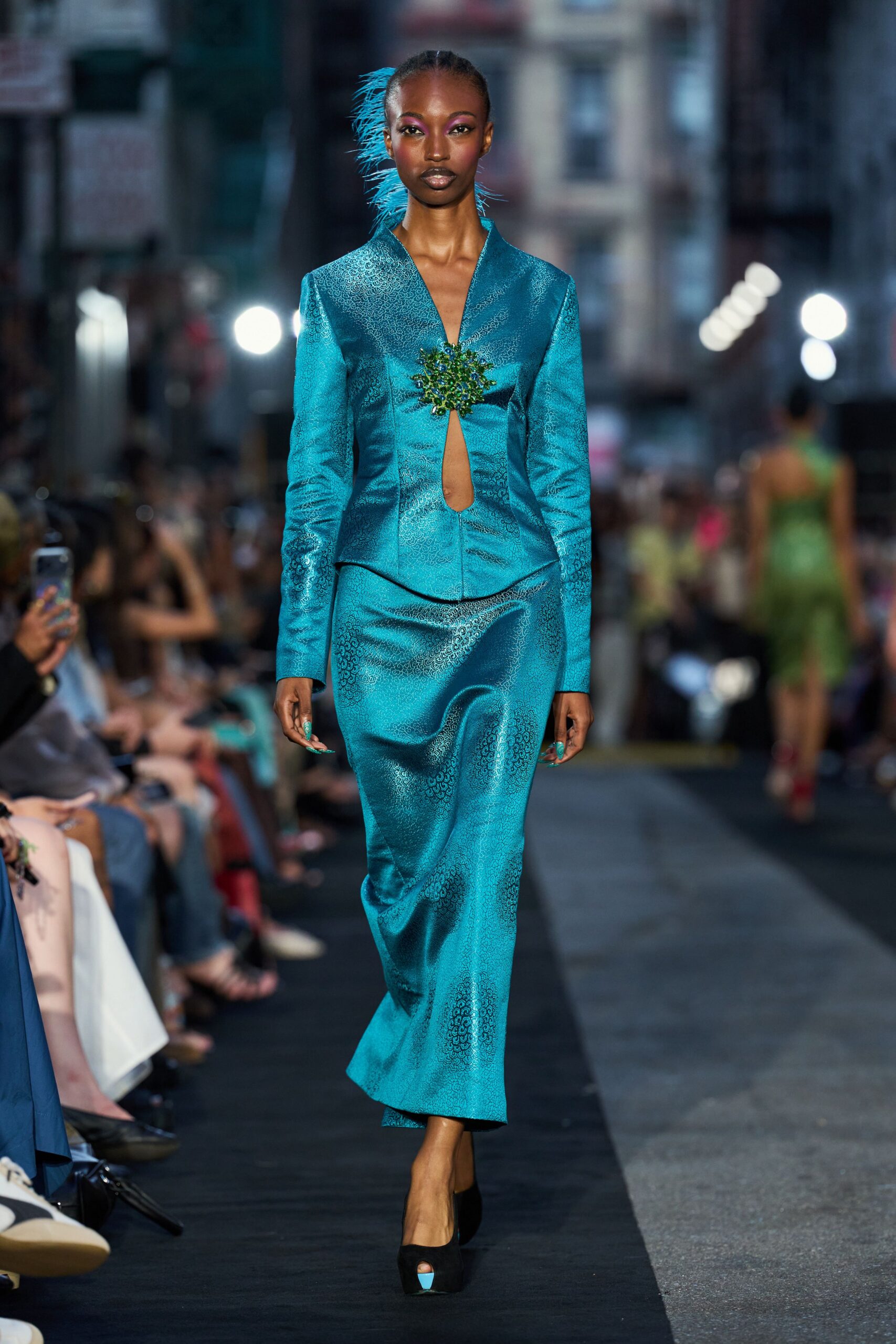 Peacock jacquard jacket with a deep slit clasped by a green jeweled burst. Matching column skirt in the same swirling jacquard, hem loosening at the ankle. Black peep-toe platforms with a flash of turquoise at the opening. Crossing downtown after rain, still carrying gala light on your clothes.