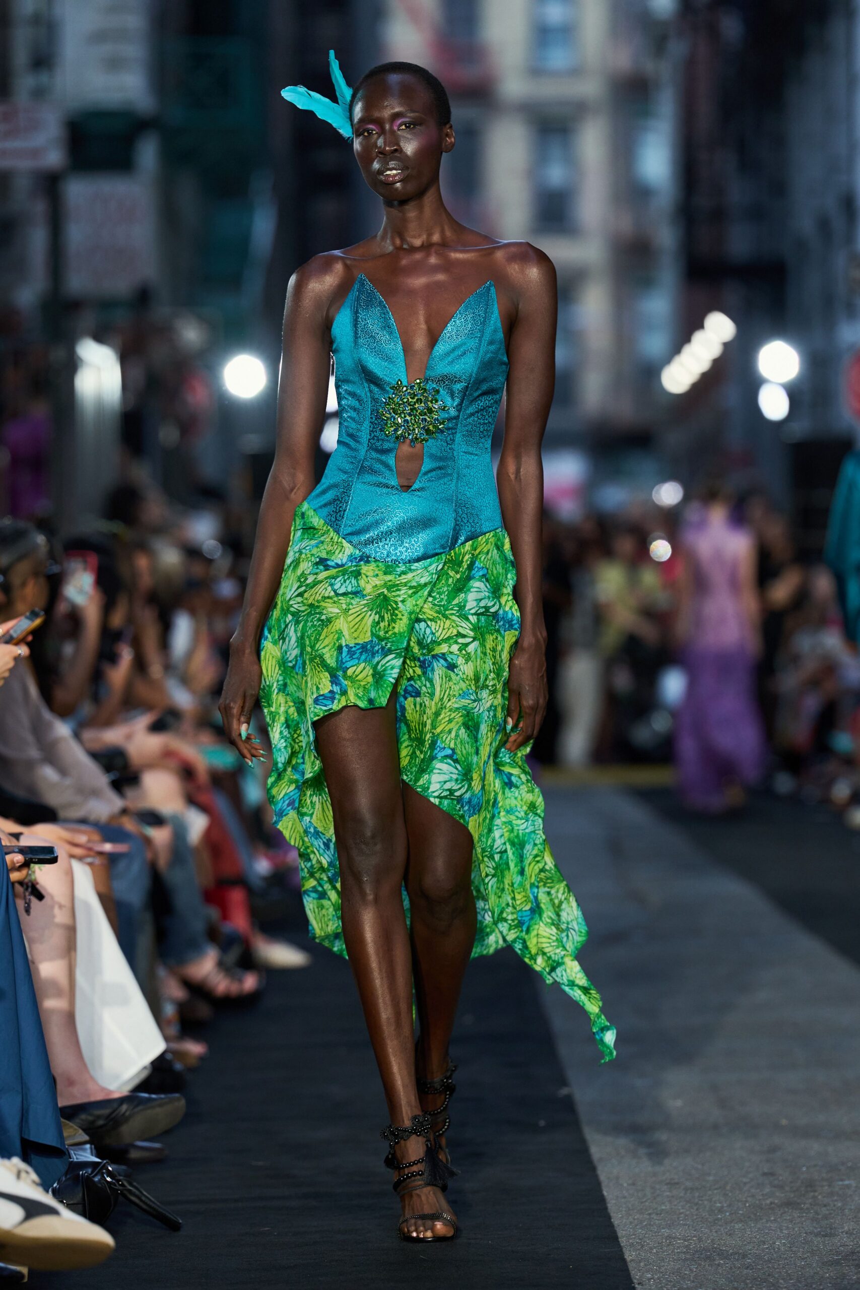 Single turquoise feather pinned upright above the temple. Turquoise jacquard bodice with a plunging notch and beaded cutout. Handkerchief skirt in lime palm print, trailing longer at one side. Black beaded lace-up sandals with a tassel at the ankle. Crossing a humid avenue just after the street fair shuts down.