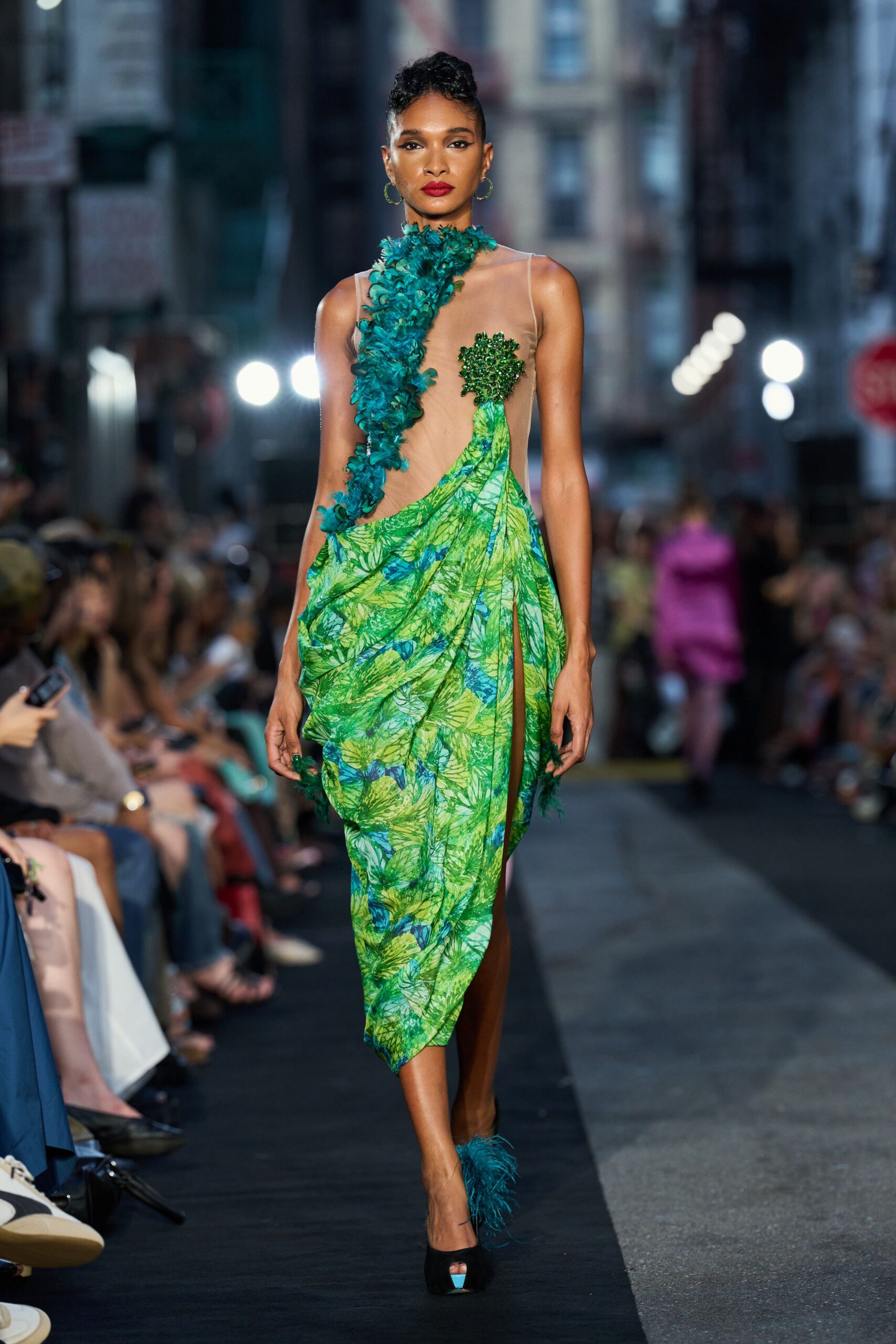 Nude illusion gown draped with lime leaf-print silk and a thigh-high slit. Teal feather spray climbing the neck, with a beaded cluster over one breast. Thin green hoop earrings. Black peep-toe heels tipped with teal feathers. Crossing a hotel lobby still damp from the fountain outside.