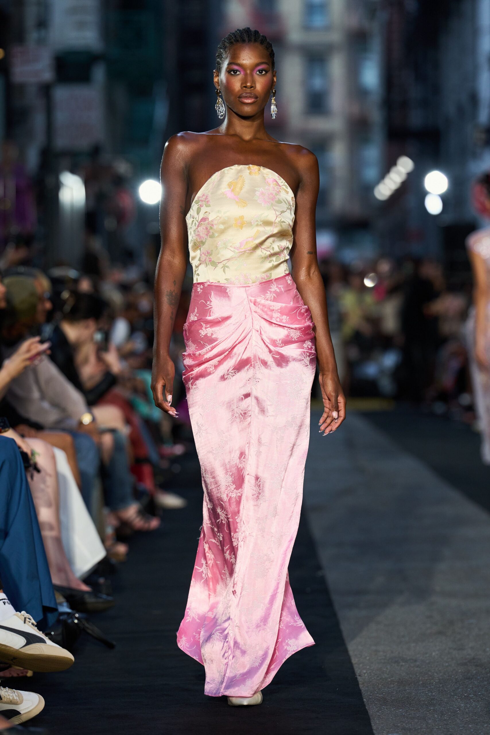 Cream satin bustier printed with washed pink blossoms and pale birds. Blush-to-rose satin column skirt, ruched into a pinched front seam. Long crystal drop earrings. Square-toe ivory pumps peeking under the hem. Crossing to the wedding after-party while the florist is still sweeping petals.