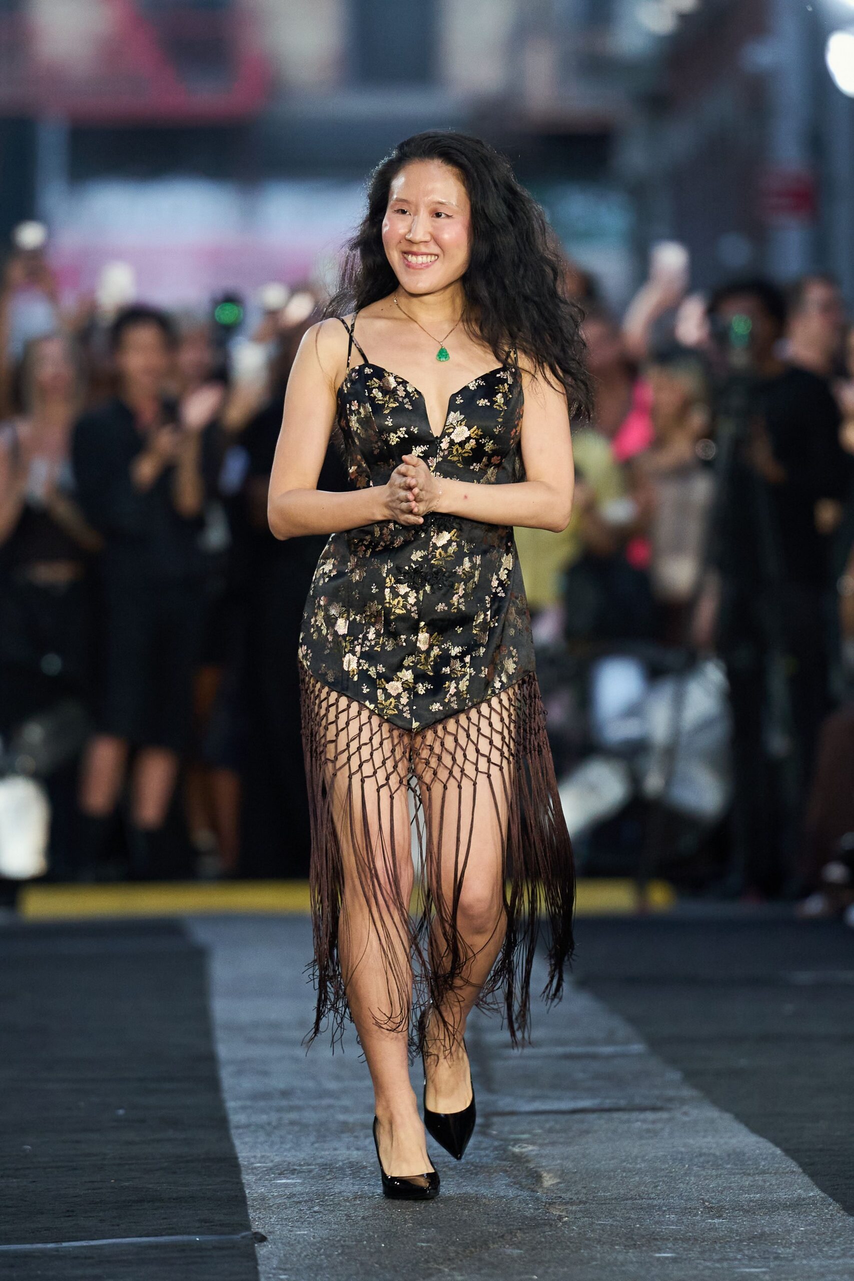 Small green teardrop pendant on a fine chain. Black floral brocade slip dress, plunge neckline and long knotted fringe skirt. Black patent pumps with narrow pointed toes. Crossing a blocked-off avenue just after the streetlights flick on.