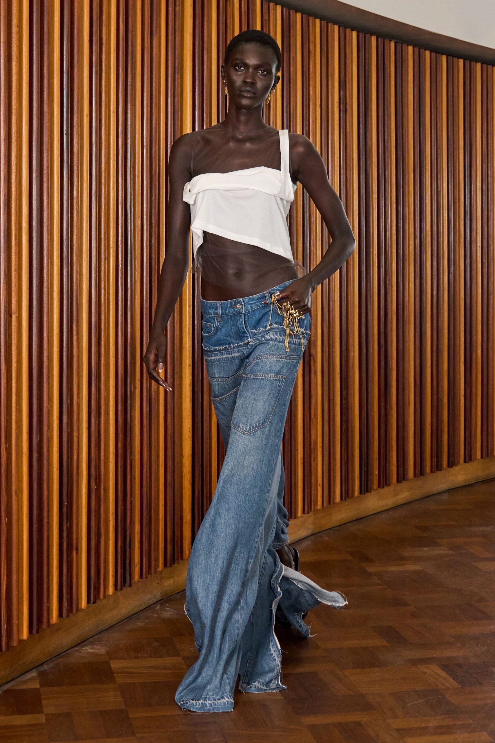 White one-shoulder drape top over sheer taupe mesh wrapping the torso. Washed blue flares with curved seam panels and a frayed trailing hem. Gold chain tassels knotted through the waistband. Slim gold drop earrings. Walking a teak-lined hotel corridor after the wedding has thinned out.