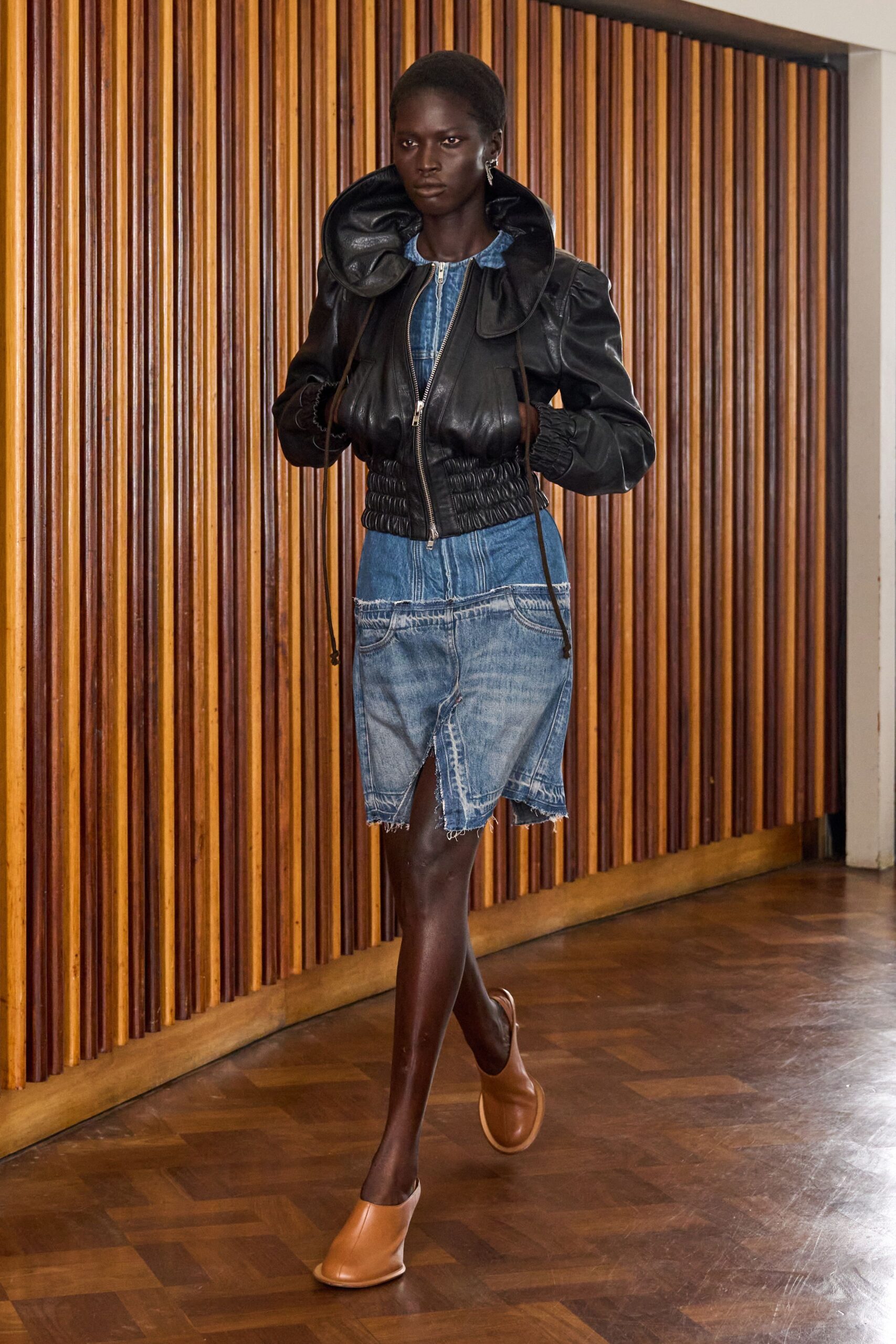 Black leather jacket with a blown-up ruff collar and smocked waist. Washed blue denim dress with dropped jean pockets and a raw split hem. Tan leather mules with rounded vamps. Walking a wood-paneled corridor before anyone opens the meeting room.