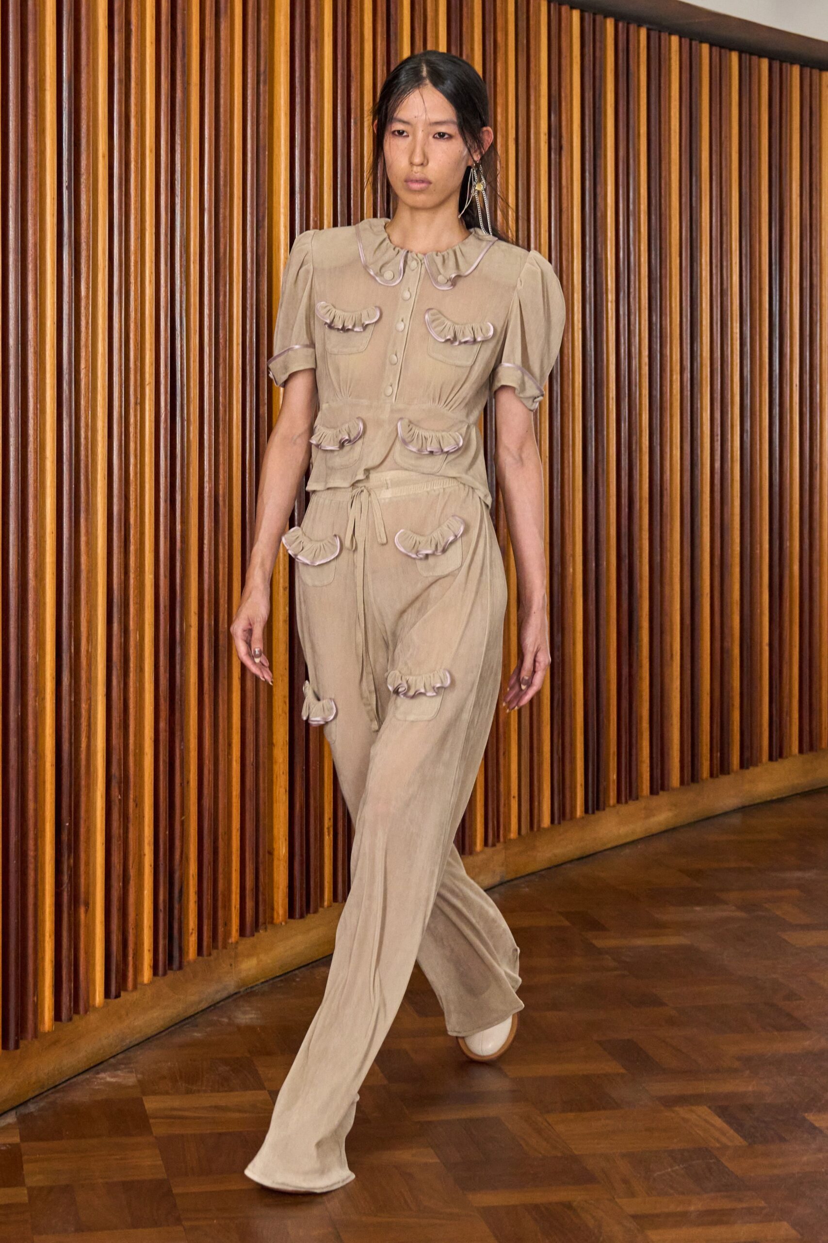 Oat voile blouse with puff sleeves and lilac-piped ruffles on every pocket. Loose matching trousers, drawstring waist, more ruffled patch pockets climbing one leg. Cream rounded flats. Walking the corridor of an old civic building after the air-conditioning failed.