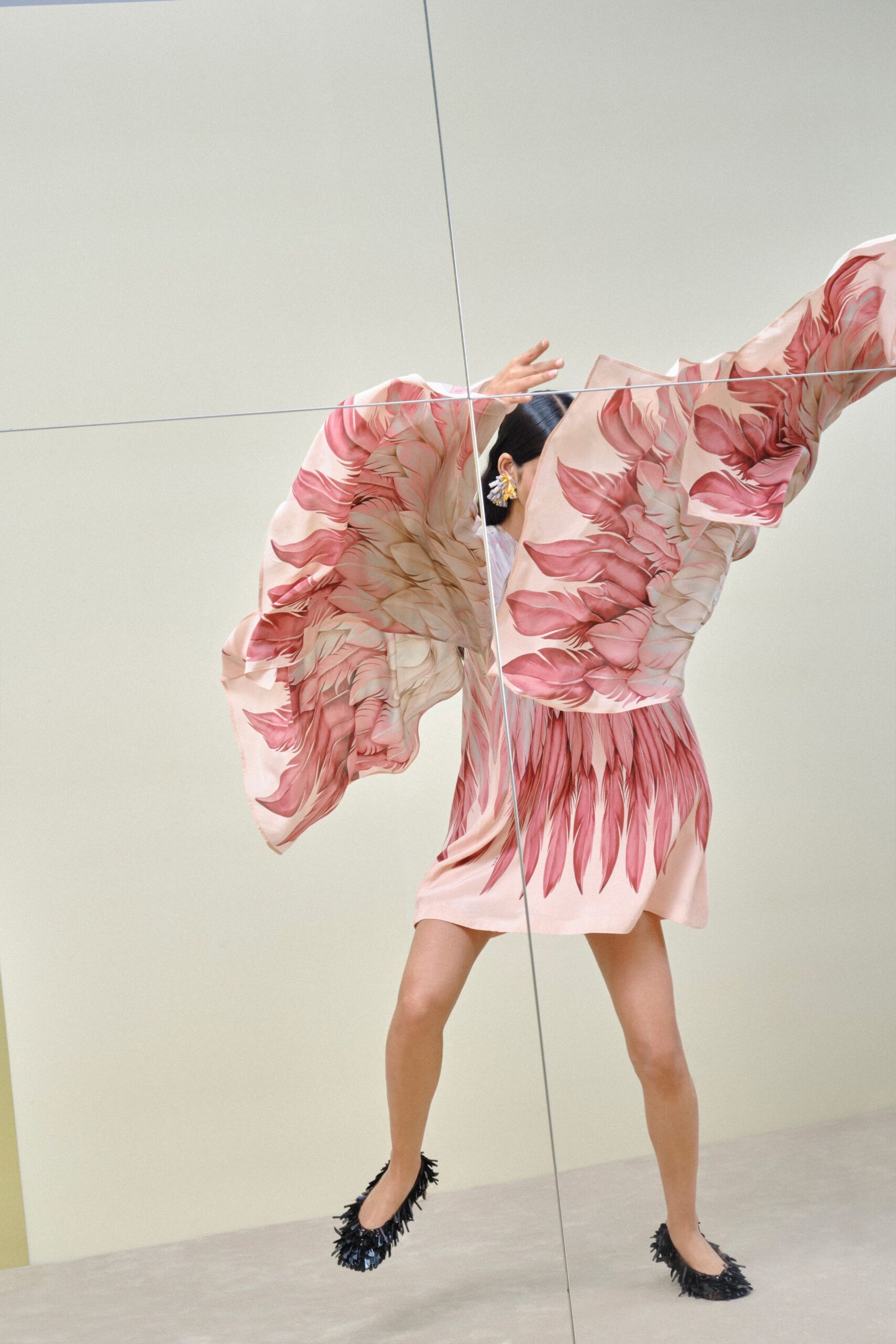 Blush satin mini dress with oversized kimono sleeves and painterly petal print. Crystal cluster earrings peeking from slicked-back hair. Black flats ringed with stiff raffia fringe. Crossing a hotel atrium while the flower delivery is still being unboxed.
