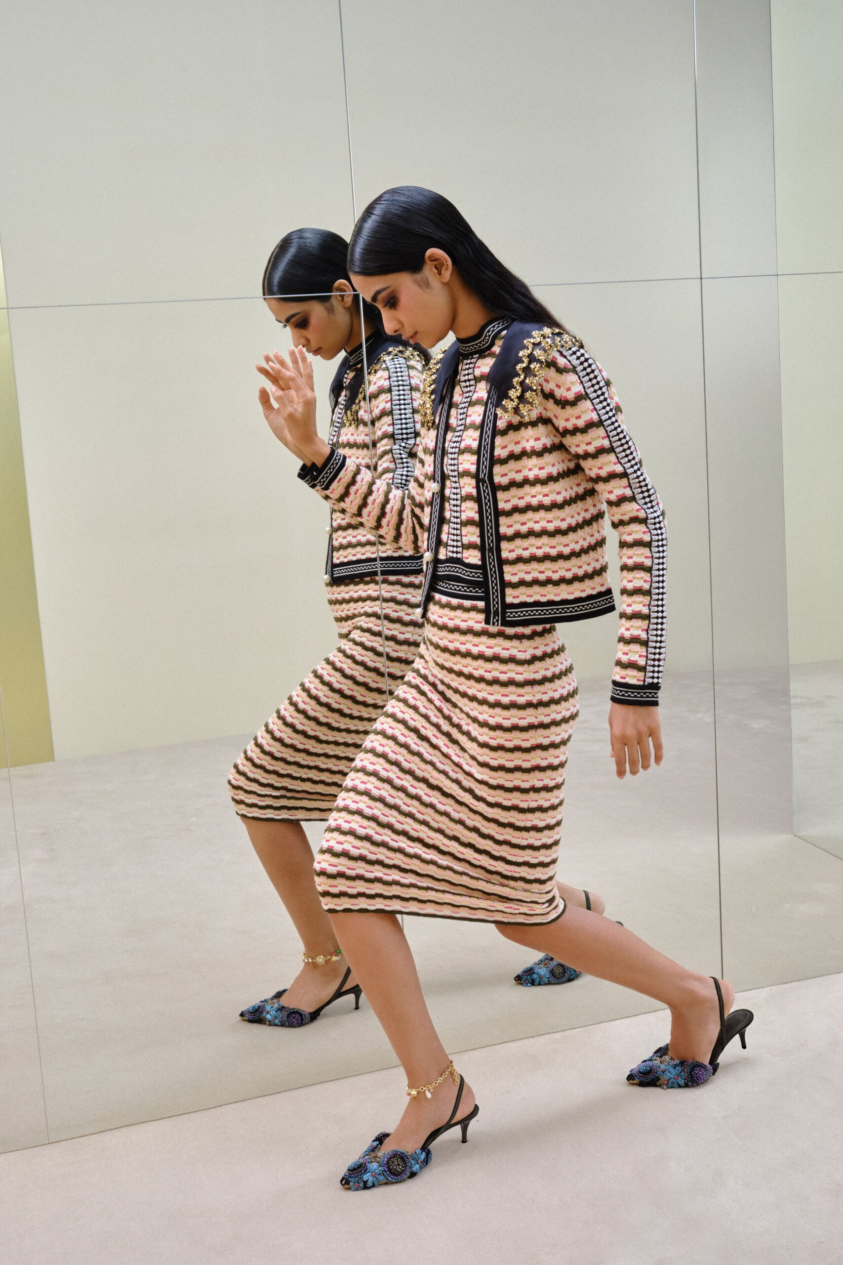 Boxy striped knit jacket with black braid borders and gold beaded shoulders. Matching ribbed knit pencil skirt, hem curving slightly off level. Gold charm anklets. Blue embroidered slingbacks with raised rosettes across the pointed toe. Crossing a hotel corridor too early for breakfast, mirrors everywhere.