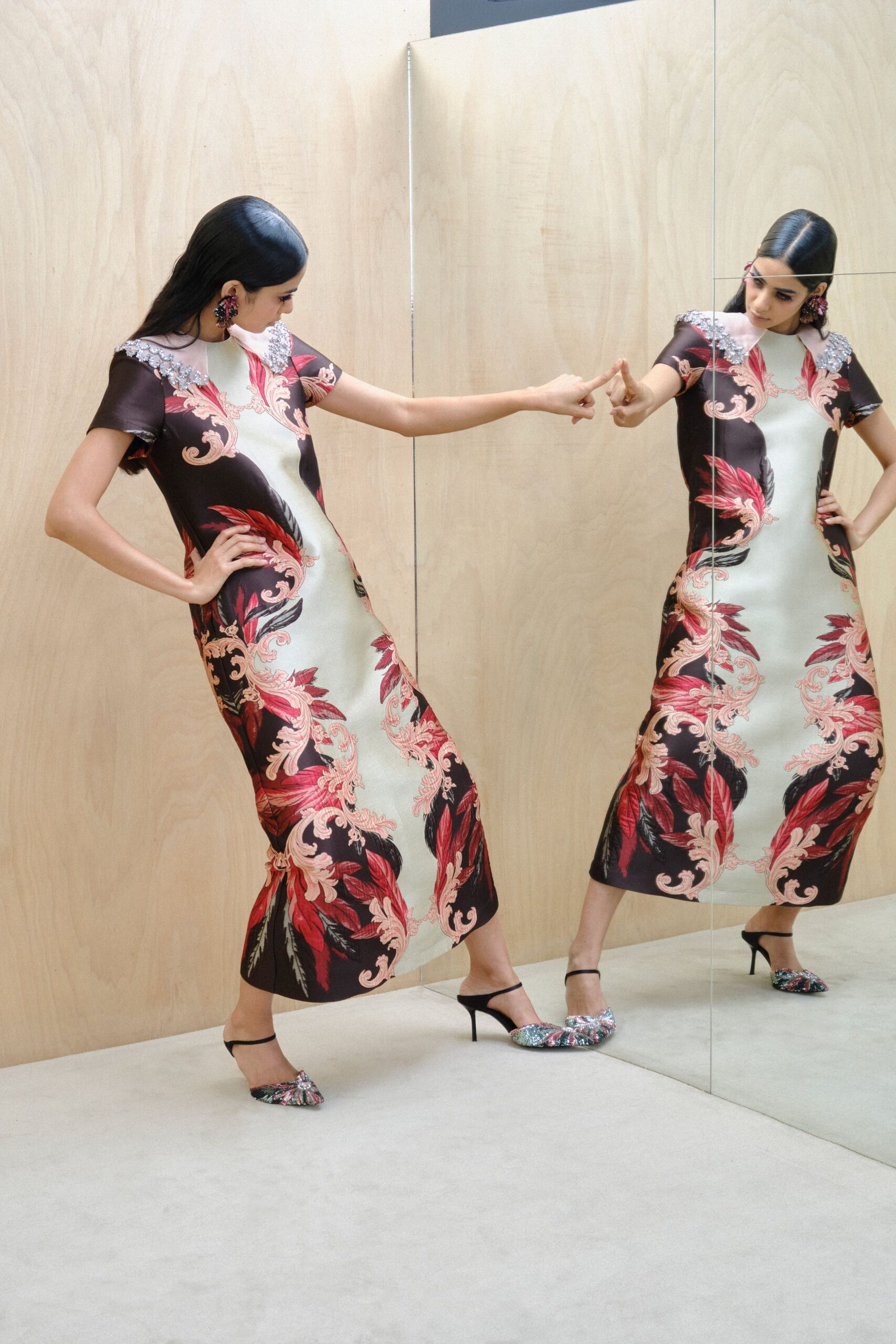 Burgundy cluster earrings with bead-heavy petals brushing the jaw. Pale celadon column dress printed with crimson baroque plumes, silver paillettes crusting the shoulders. Black slingback mules with a glittered ruched vamp. Touching your reflection in a hotel corridor before the wedding lunch.