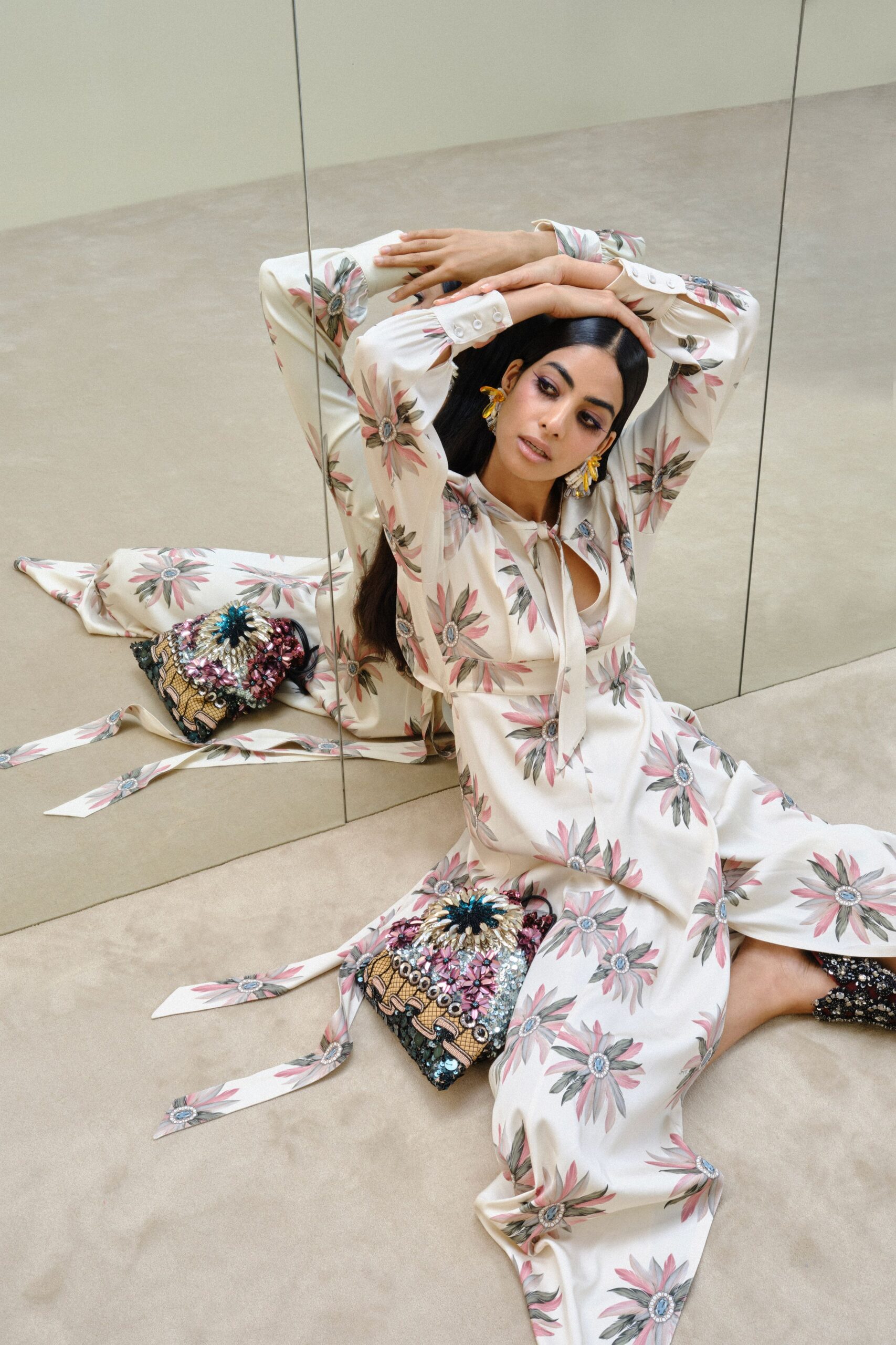 Ivory satin dress scattered with pink daisy bursts, neck ties trailing on the floor. Amber and crystal floral drops brushing the jaw. Beaded pouch packed with sequins and raffia petals. Black ankle boots crusted with dense paillettes. Afternoon in a mirrored fitting room, one shoe half off already.