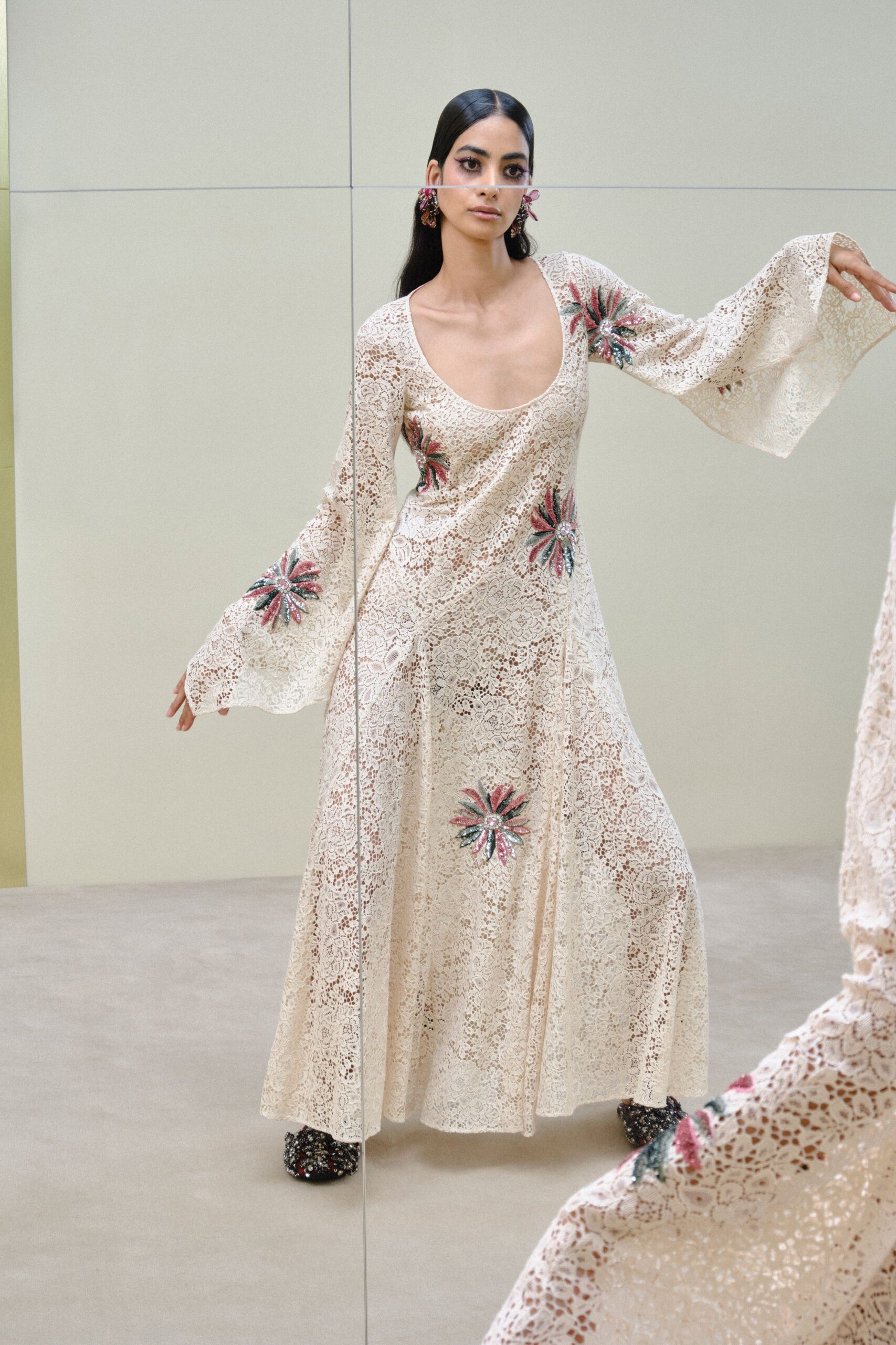 Pink crystal flower clusters brushing the jaw. Ivory openwork lace gown with a deep scoop neck and flared cuffs. Beaded pink starbursts scattered at sleeve, hip and hem. Black floral brocade mules, densely jeweled across the vamp. Hotel atrium wedding, half an hour before anyone finds the bride.