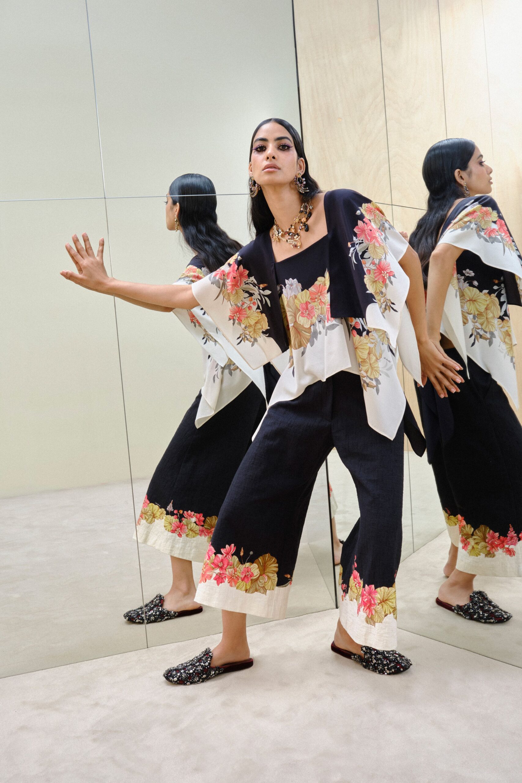 Black draped cape top with ivory panels printed in coral hibiscus. Square-neck black jumpsuit, cropped wide legs banded with the same border print. Beaded floral mules with a slight upturned toe. Walking the echoing corridor outside a hotel ballroom after the wedding band starts.
