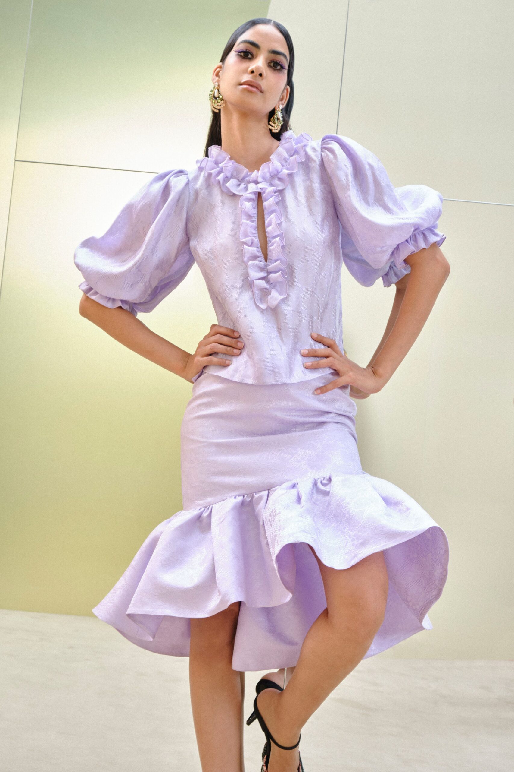 Tiered gold filigree earrings with a teardrop pearl at the center. Lilac jacquard blouse with puff sleeves and ruffles framing a narrow slit. Matching satin-jacquard skirt, fitted through the thigh then kicked into a deep flounce. Black ankle-strap heels. Garden wedding reception, the cake already leaning in the heat.